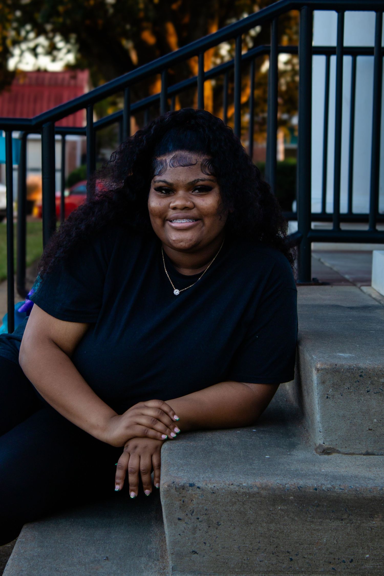 Young Black woman in black t-shirt smiles while relaxing on outdoor concrete steps at dusk.