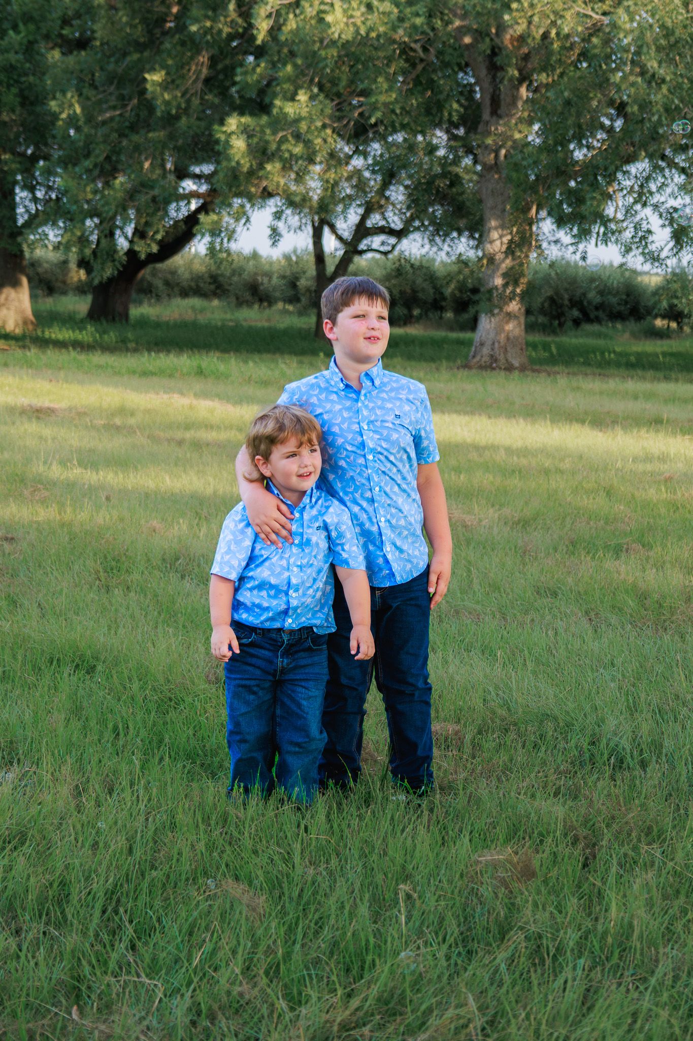 Two boys in matching blue floral shirts and jeans stand together in a green field with trees in the background.