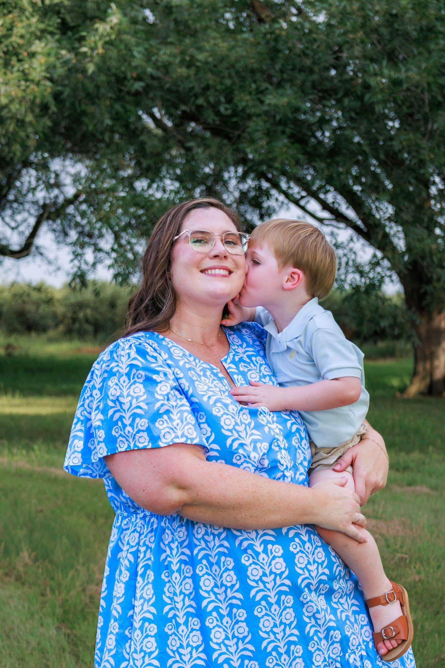 A smiling woman in a blue floral dress holds a toddler boy who gives her a sweet kiss on the cheek outdoors.