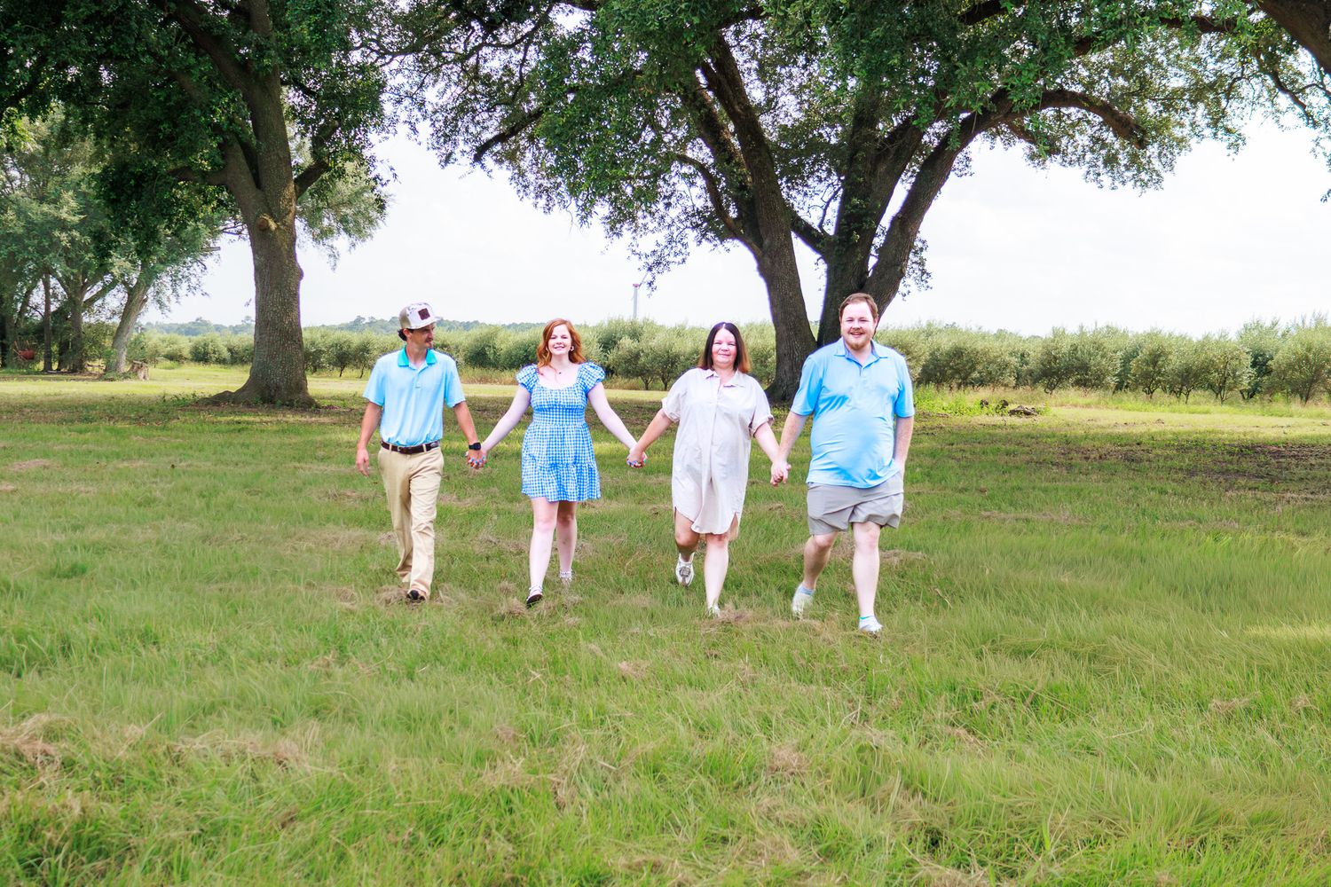 Four people holding hands walking through a lush green field with large trees on a sunny day.