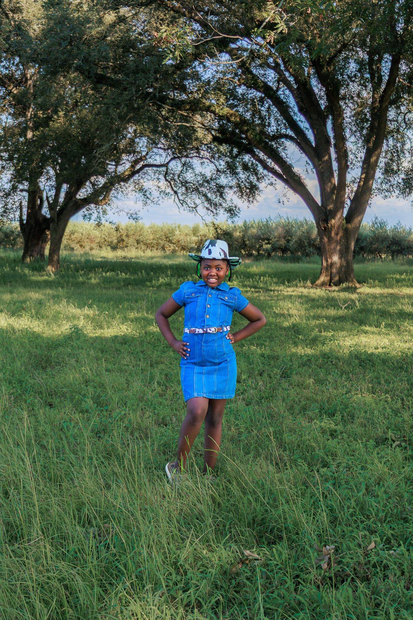 A young African woman in traditional blue attire with headpiece stands confidently in a lush green field with trees.