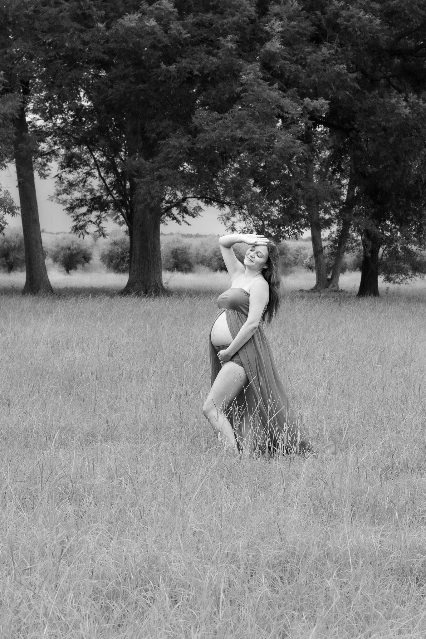 Black and white photo of a pregnant woman in a field wearing a flowing skirt, posing gracefully among tall grass and trees.