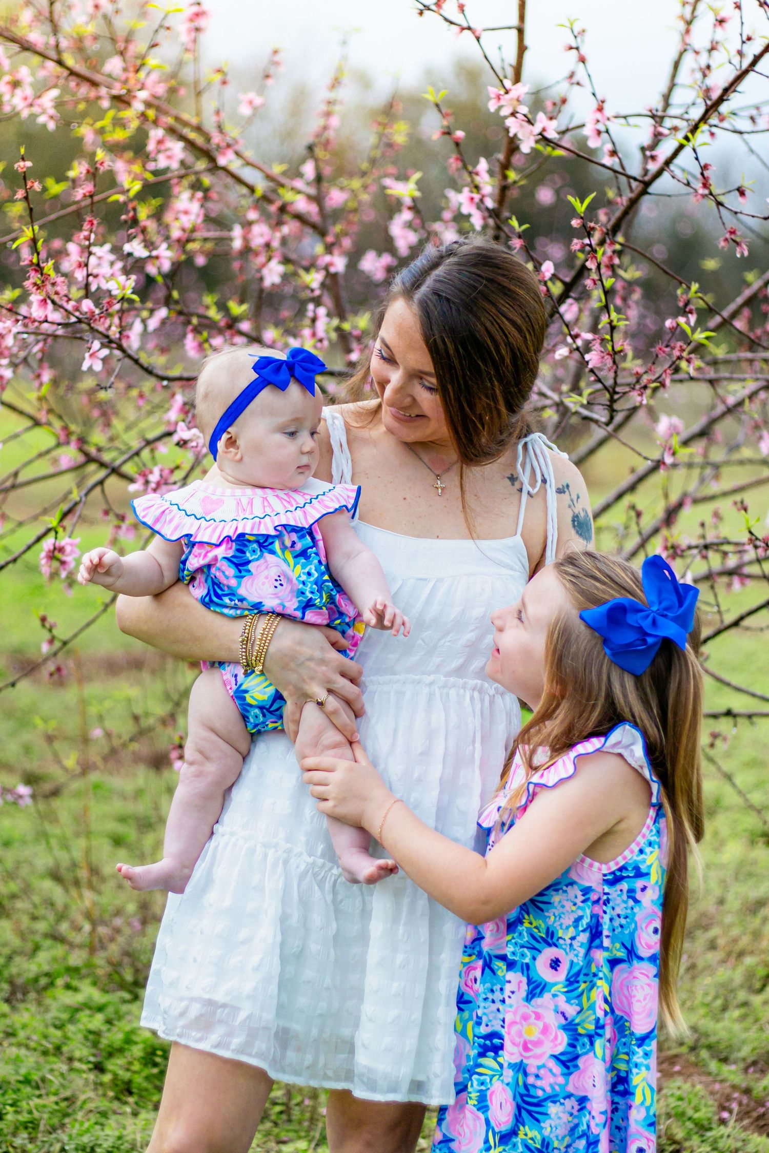 Mother in white dress holds infant while older daughter in blue floral dress stands beside her.