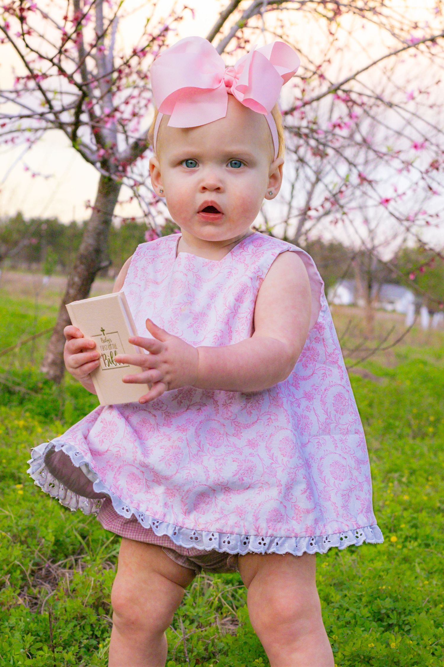 Blue-eyed baby girl in pink floral dress and bow headband holding small book outdoors among spring trees.