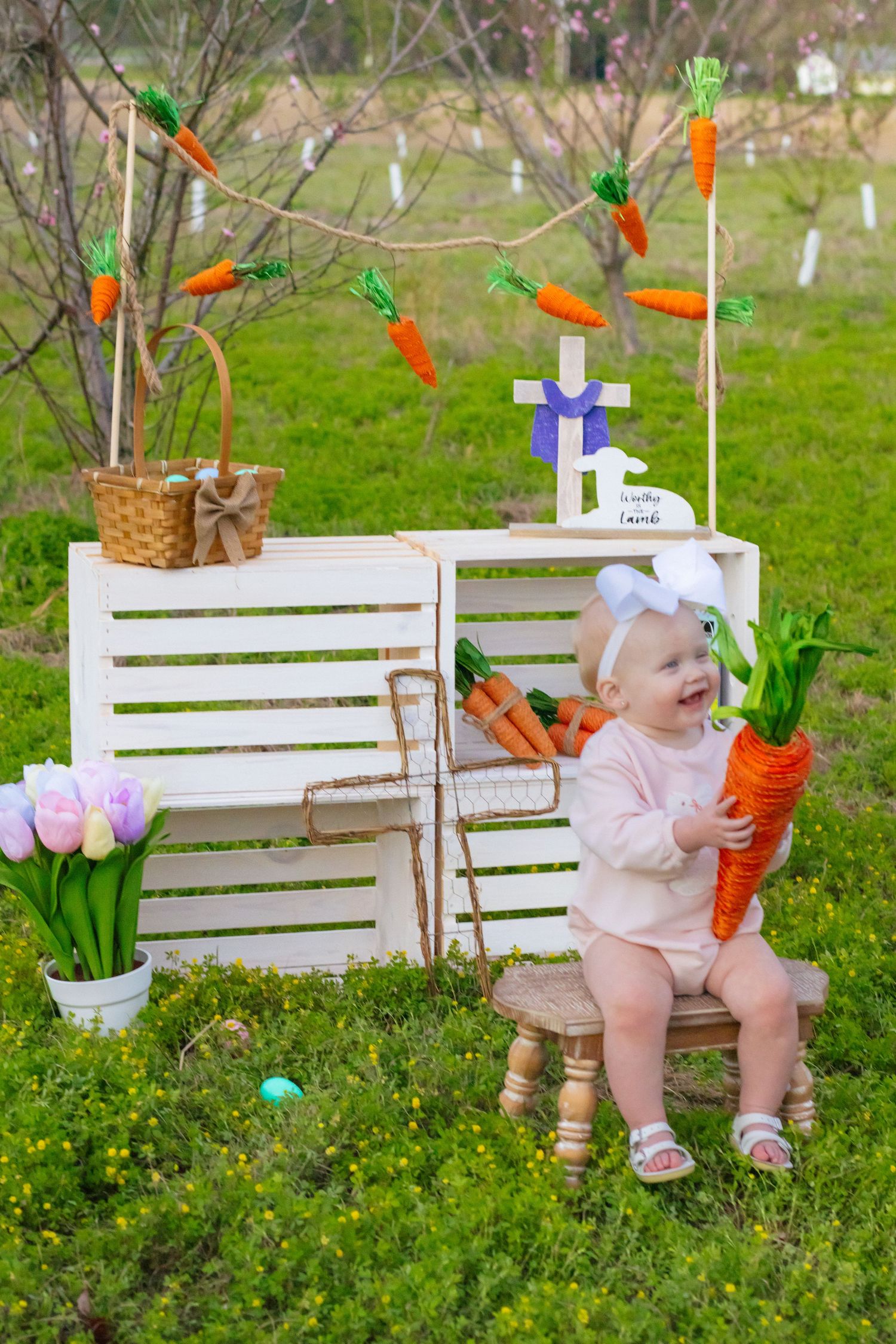 Happy baby with bunny ears sits on bench holding decorative carrot at Easter setup.