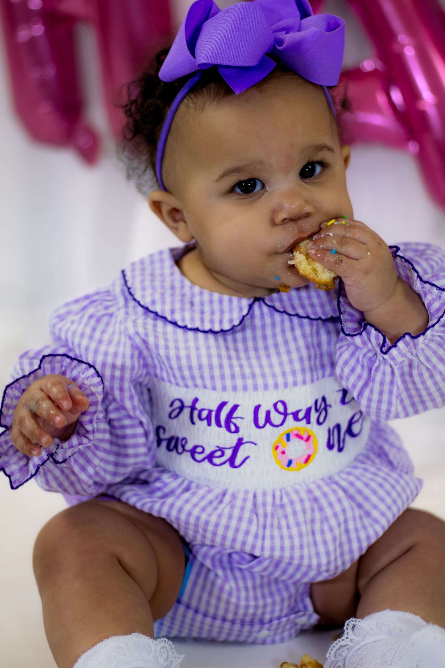 Close-up of baby in Half Way 2 Sweet One purple romper happily eating a donut.