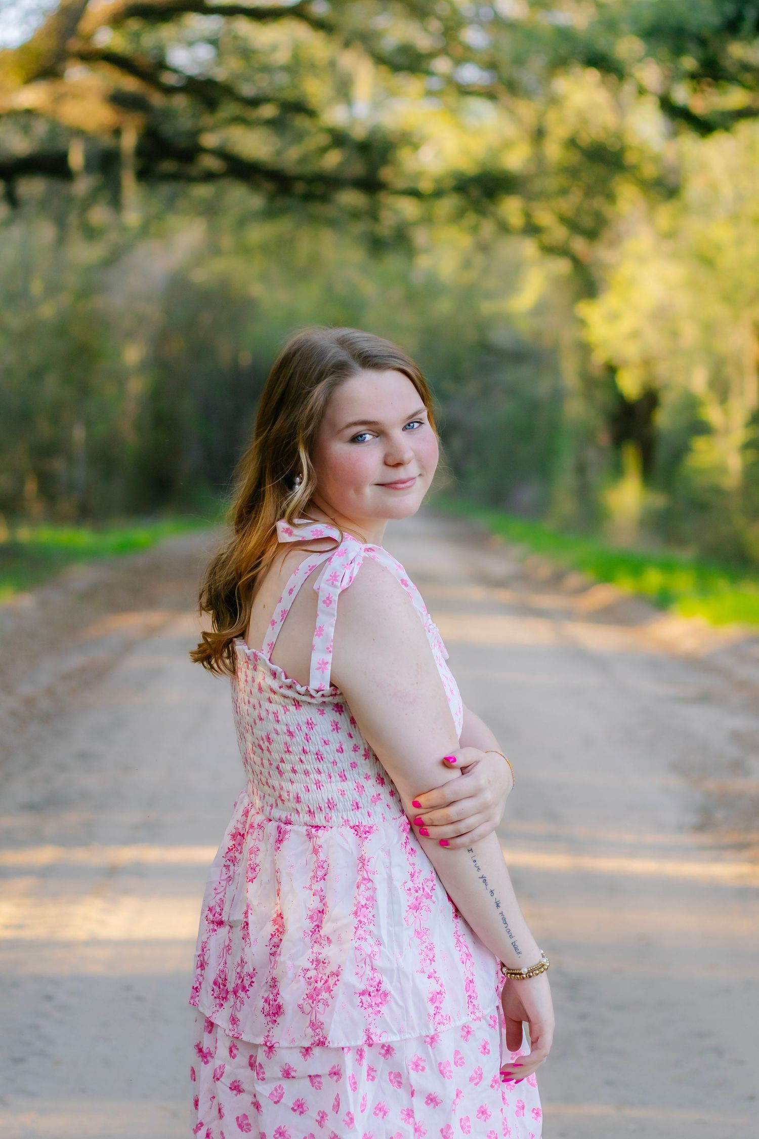 Young girl in a pink floral tiered dress poses on a scenic tree-lined dirt road.