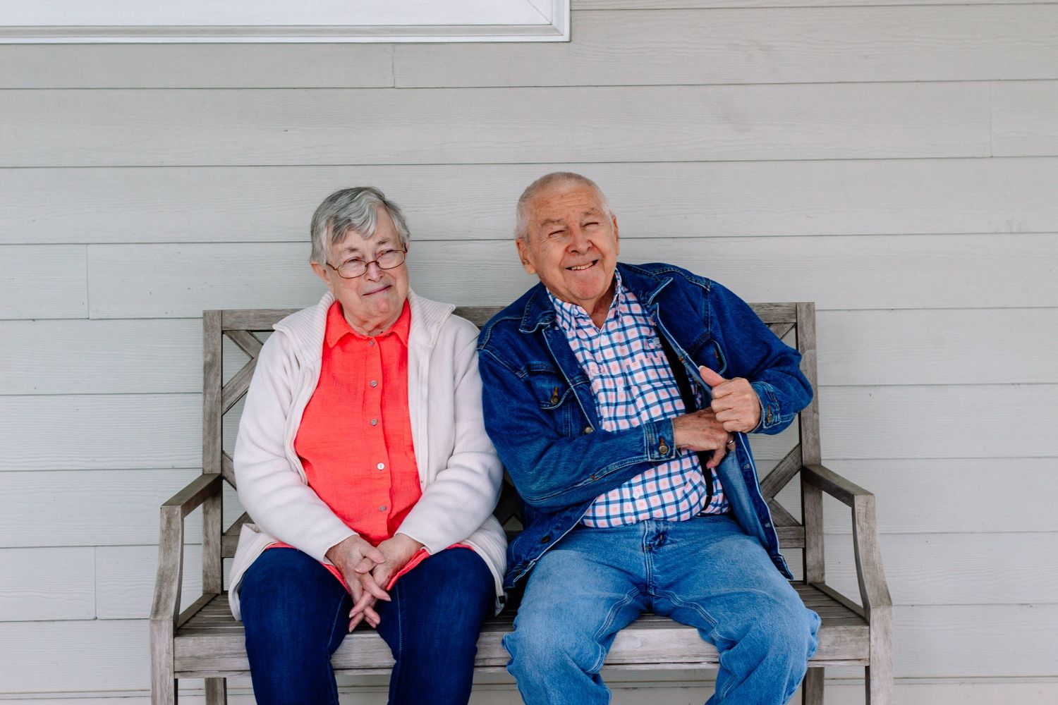 Elderly couple smiling and sitting together on a wooden bench on a porch.