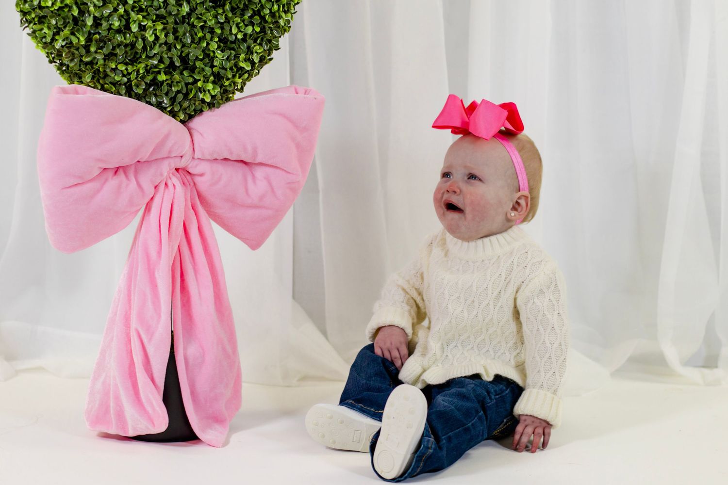 Baby girl in cream sweater and pink bow headband sits on white floor beside a large pink decorative bow.