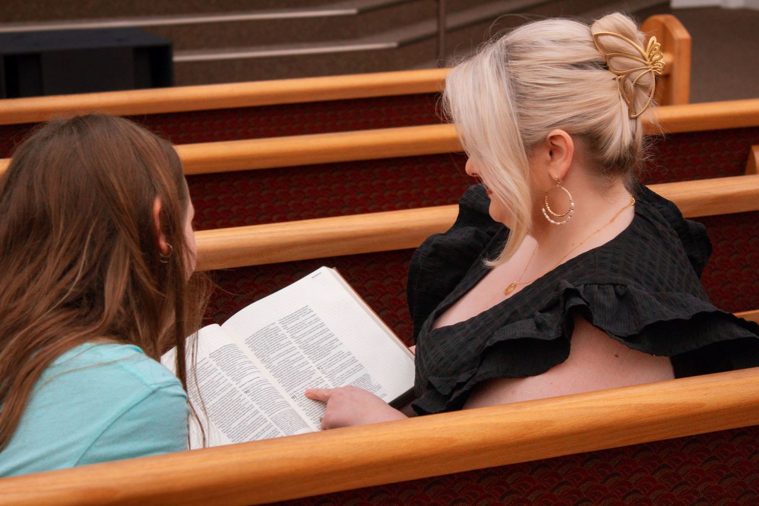 Two women sit together in church pews reading an open Bible near a stained glass window.