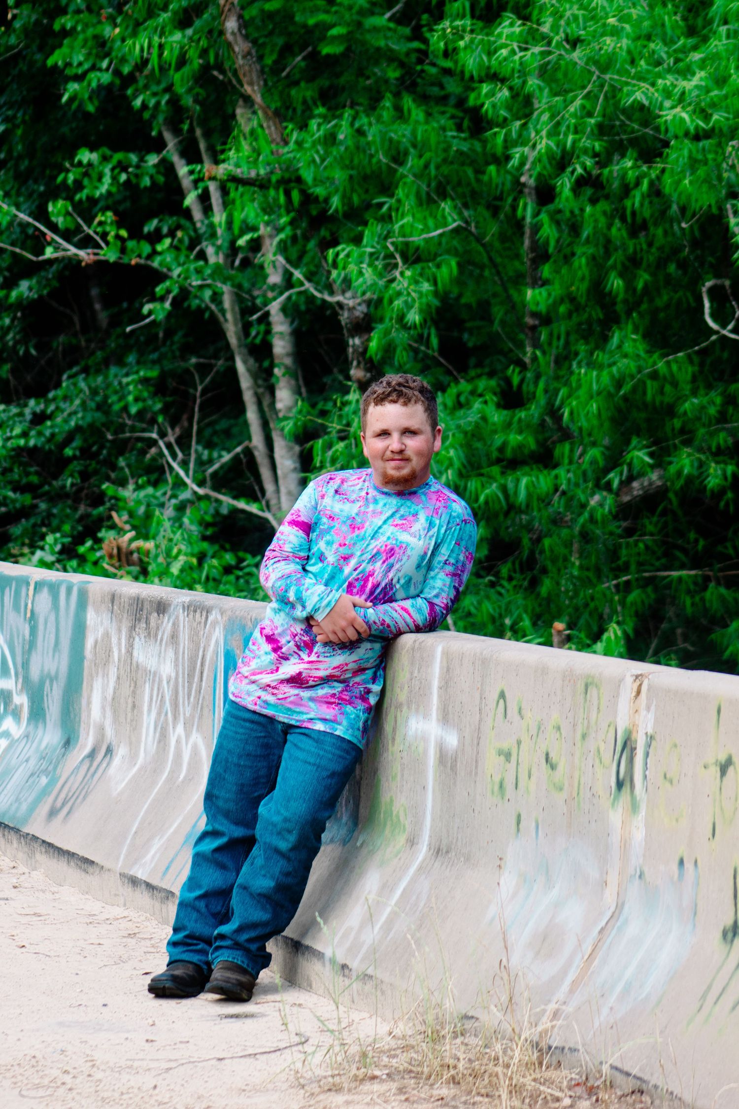 Man in tie-dye long sleeve shirt leaning against a graffiti concrete bridge wall.