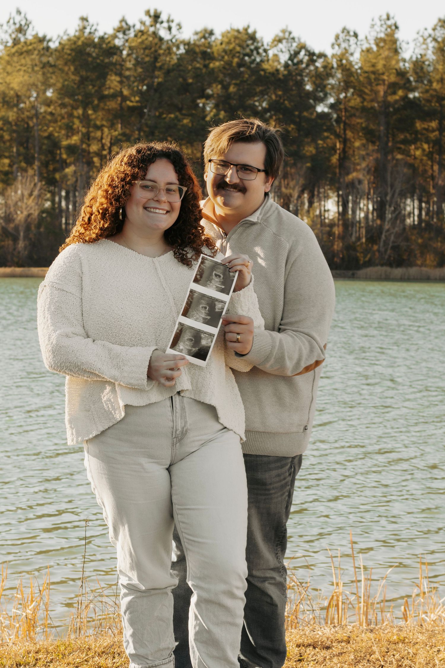 Couple smiling and holding ultrasound photos by a lake in autumn.