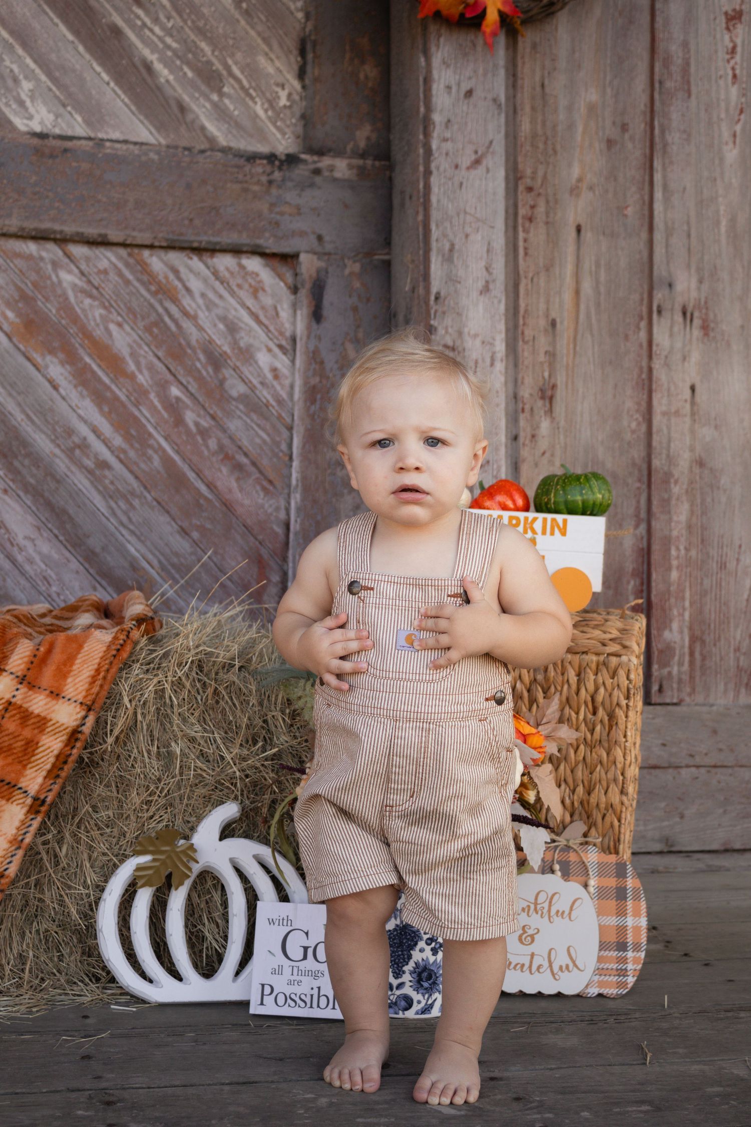Toddler boy in tan overalls stands barefoot near hay bales and autumn harvest decorations at a barn.