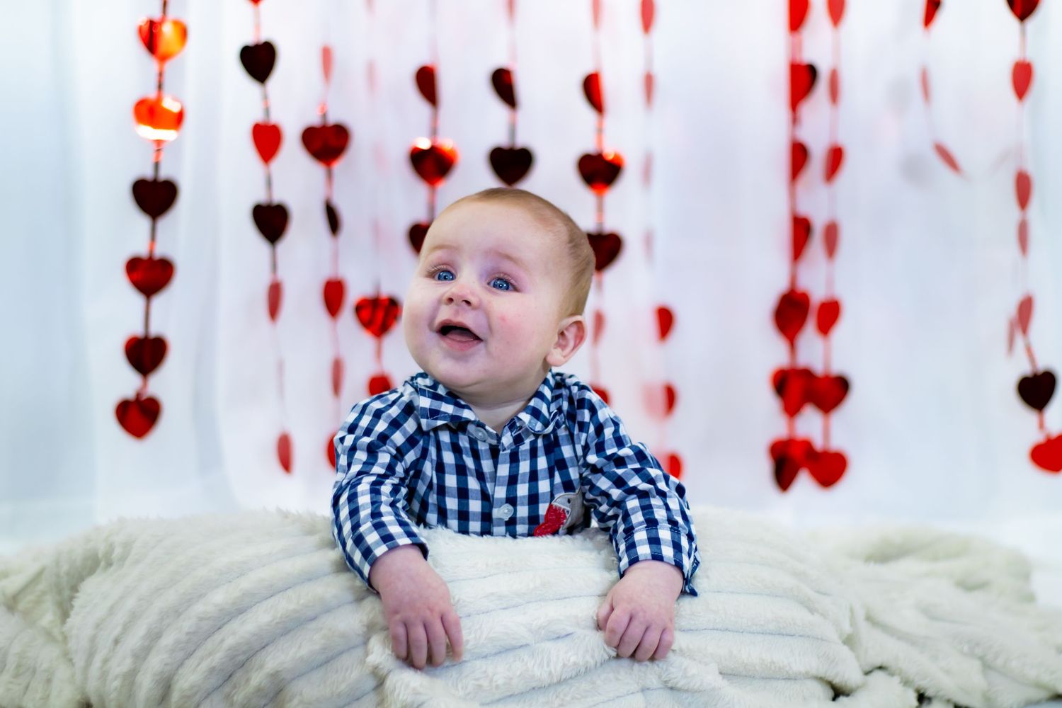 Adorable baby in plaid shirt lies on white blanket surrounded by hanging red heart garland decor.