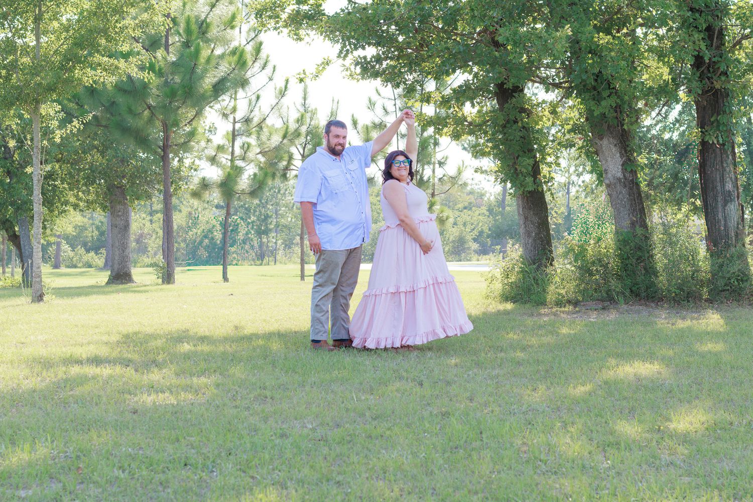 Pregnant couple holds hands while posing together in a sunny green park for maternity photos.