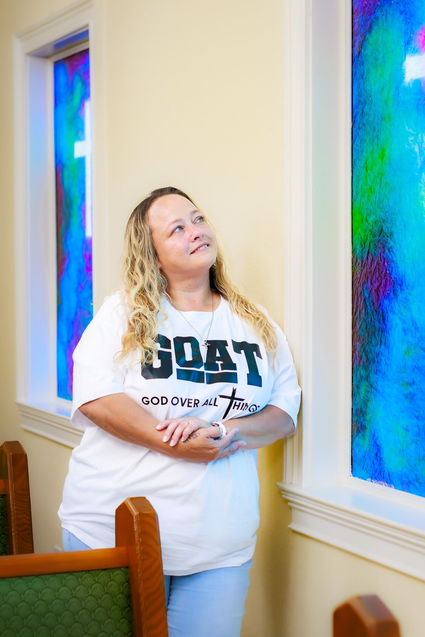 Woman wearing GOAT God Over All Things shirt looking upward near vibrant stained glass church windows.