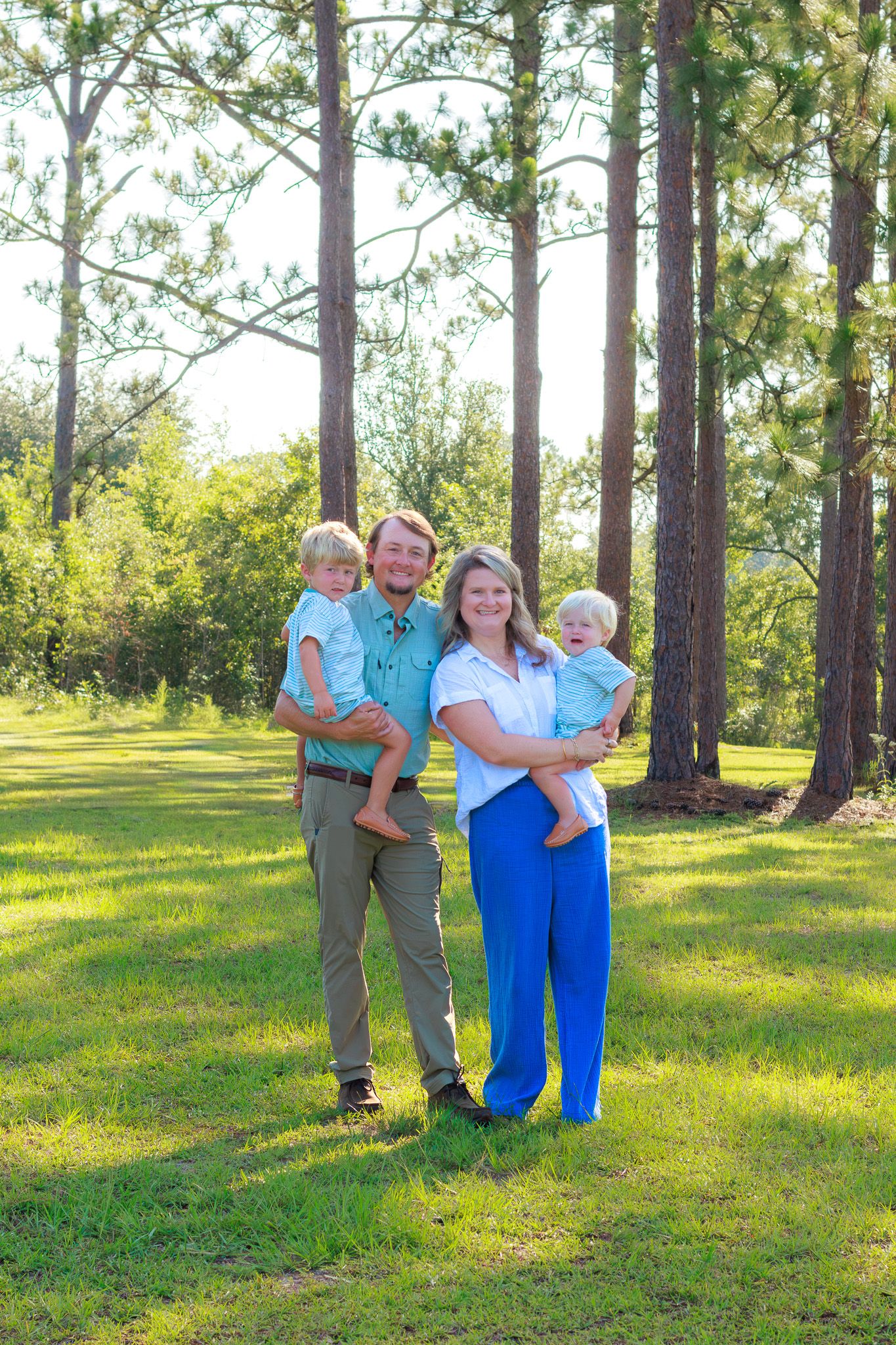 A happy family of four poses outdoors on green grass surrounded by tall pine trees on a sunny day.