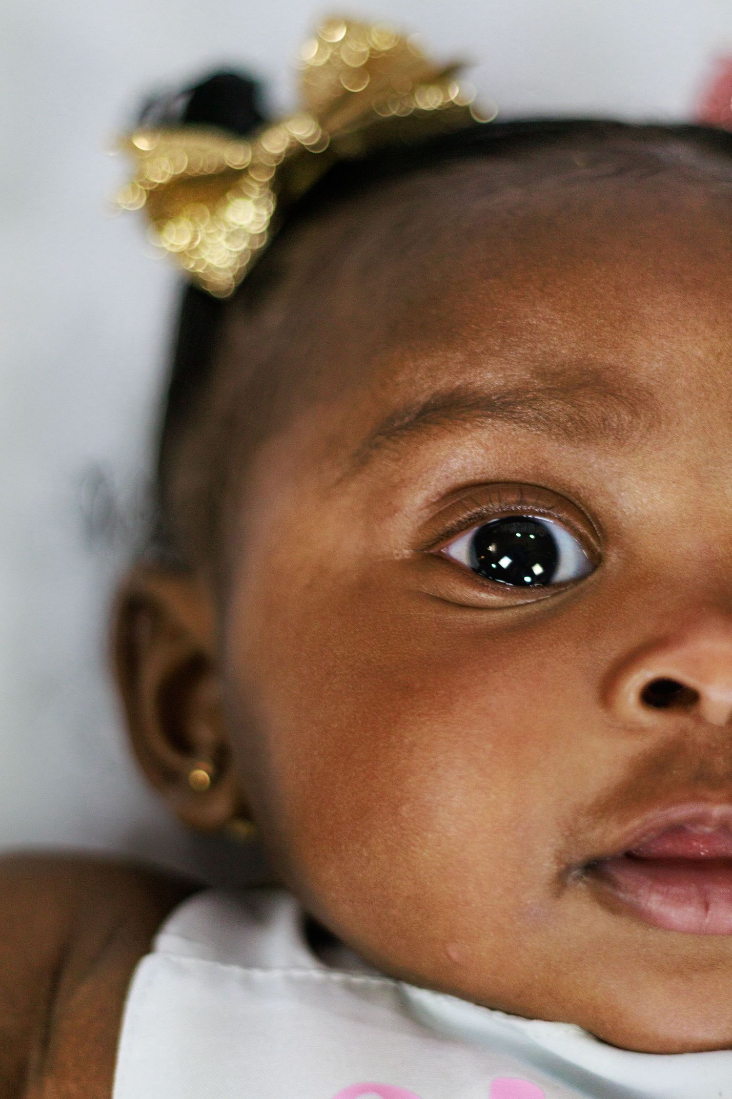 Close-up portrait of a baby girl with a gold bow headband and gold earring, looking up with wide brown eyes.