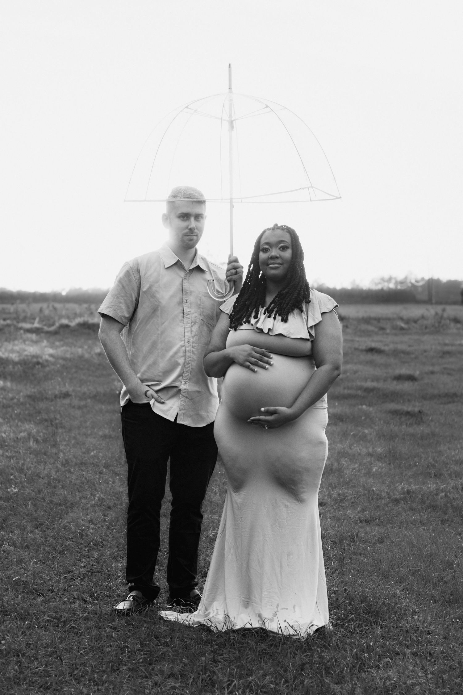 Black and white maternity photo of couple holding clear umbrella in field, pregnant woman in flowing dress cradling baby bump.