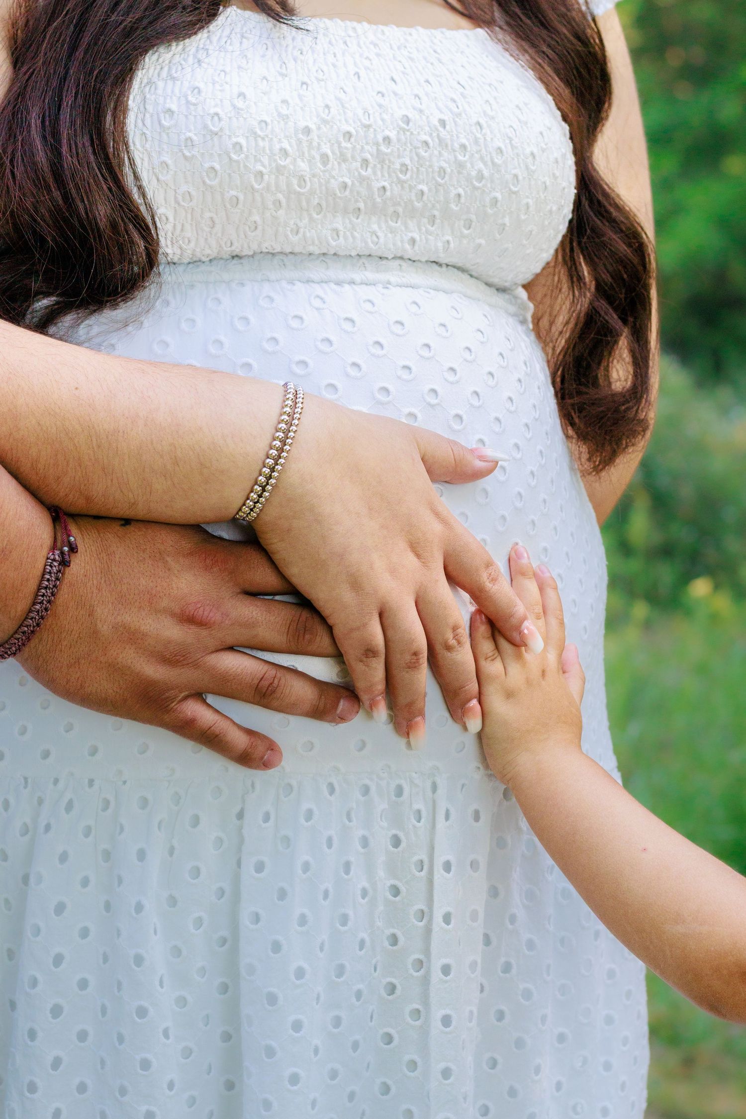 Close-up of pregnant womans baby bump with husbands and childs hands resting on white dress.