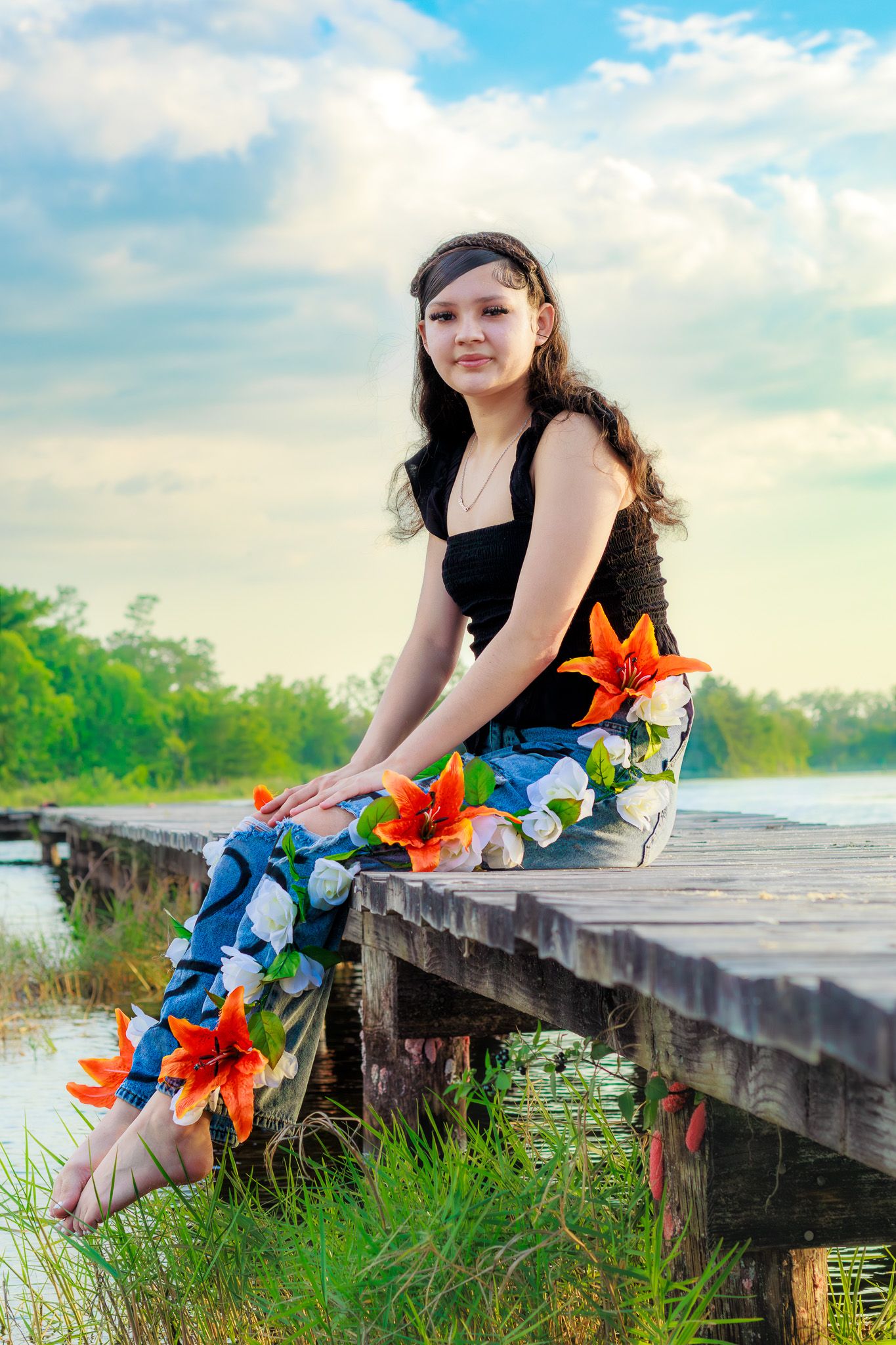 Young woman sitting on a wooden dock wearing floral pants, with a serene lake and cloudy sky in the background.