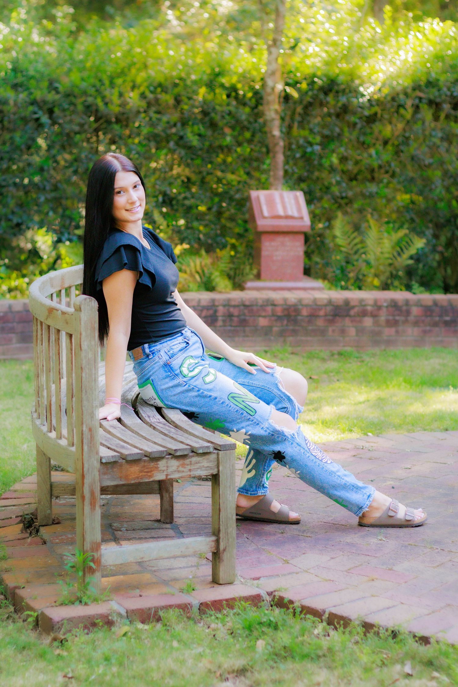 Smiling young woman with long dark hair sits on curved wooden bench in sunny garden wearing black top and painted jeans.