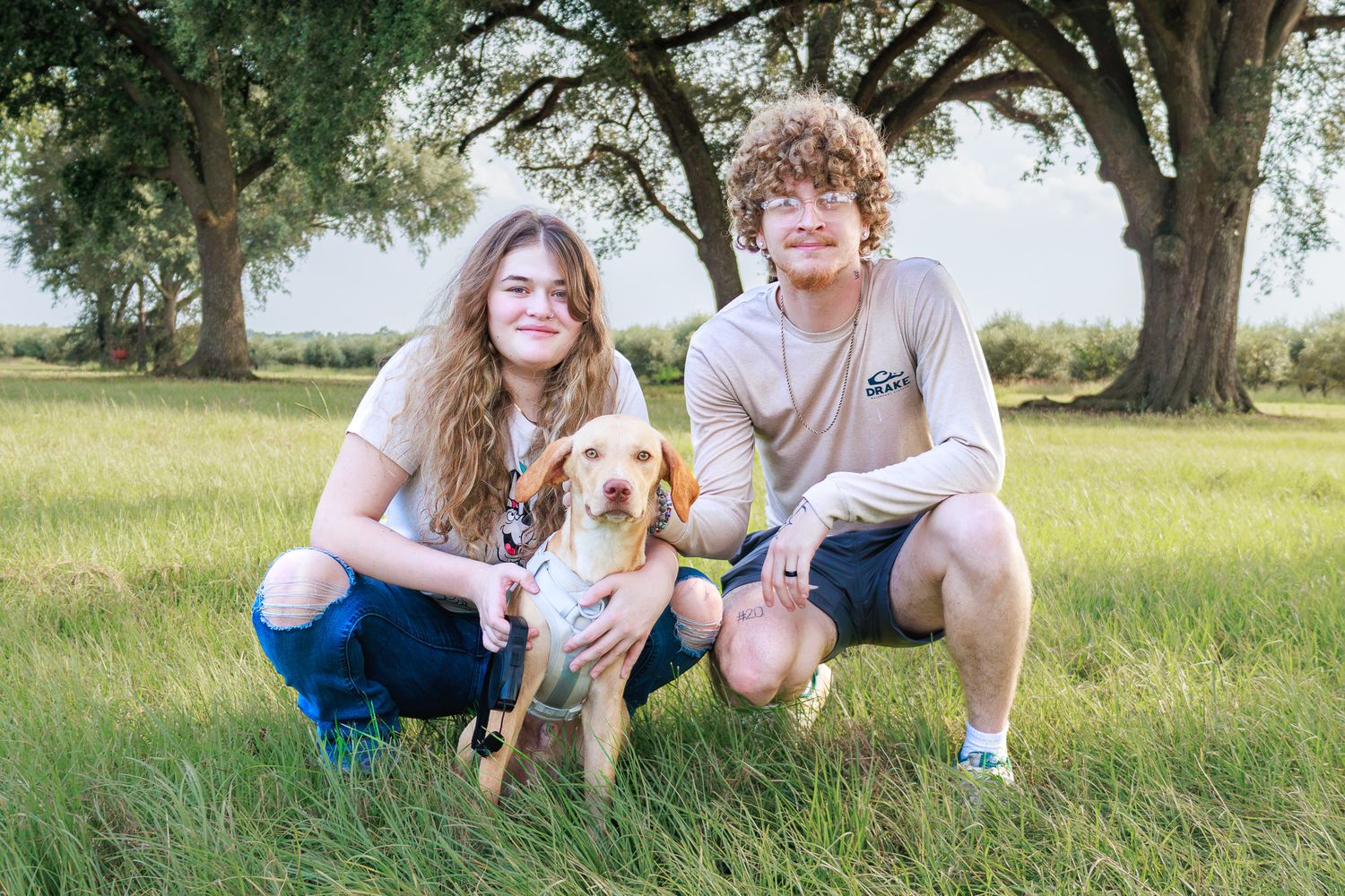 Two teens kneel with a yellow Labrador puppy in a green field under large trees on a sunny day.