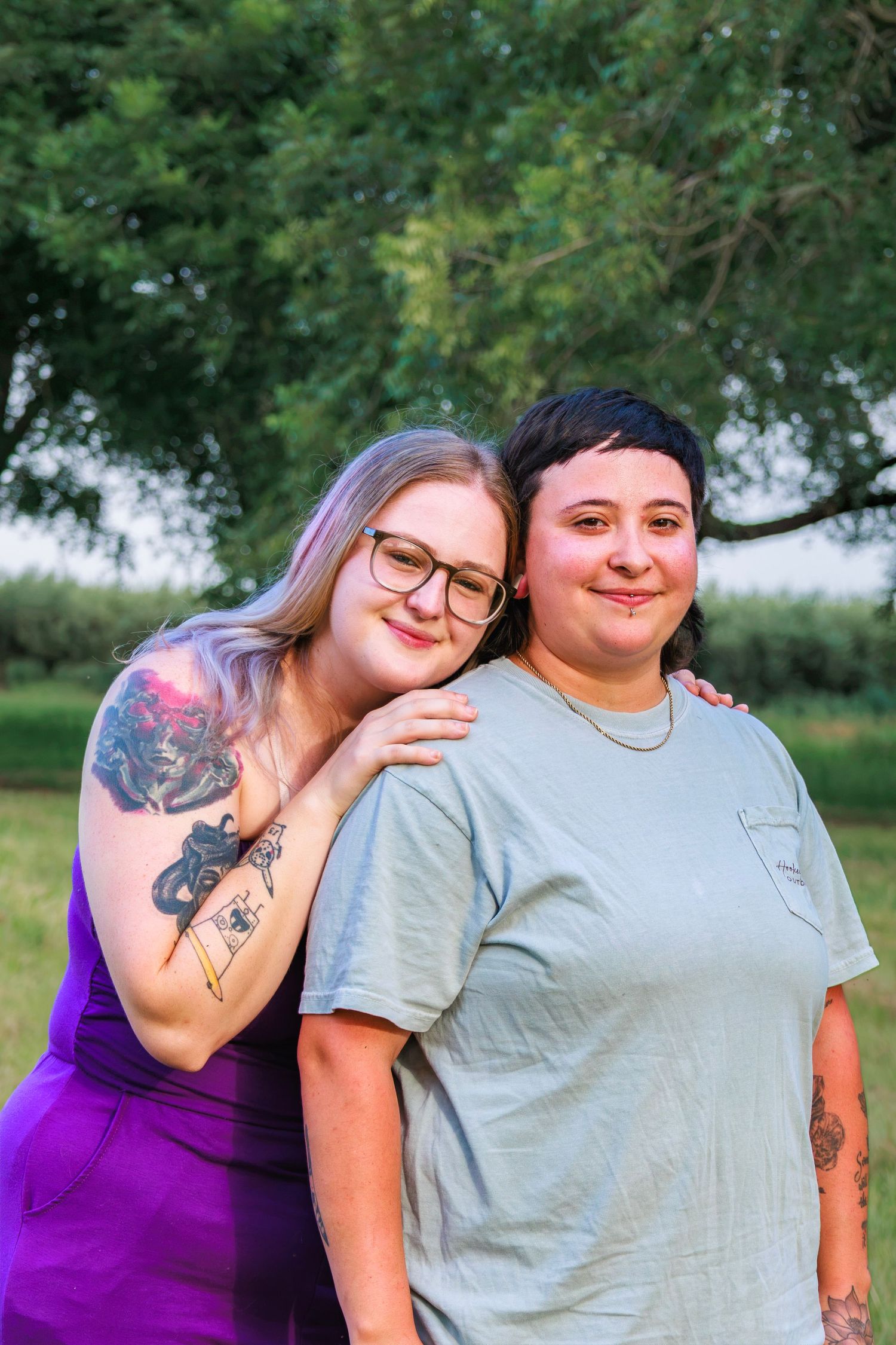 Two women smiling and posing together outdoors, one in a purple dress with tattoos, the other in a gray t-shirt.