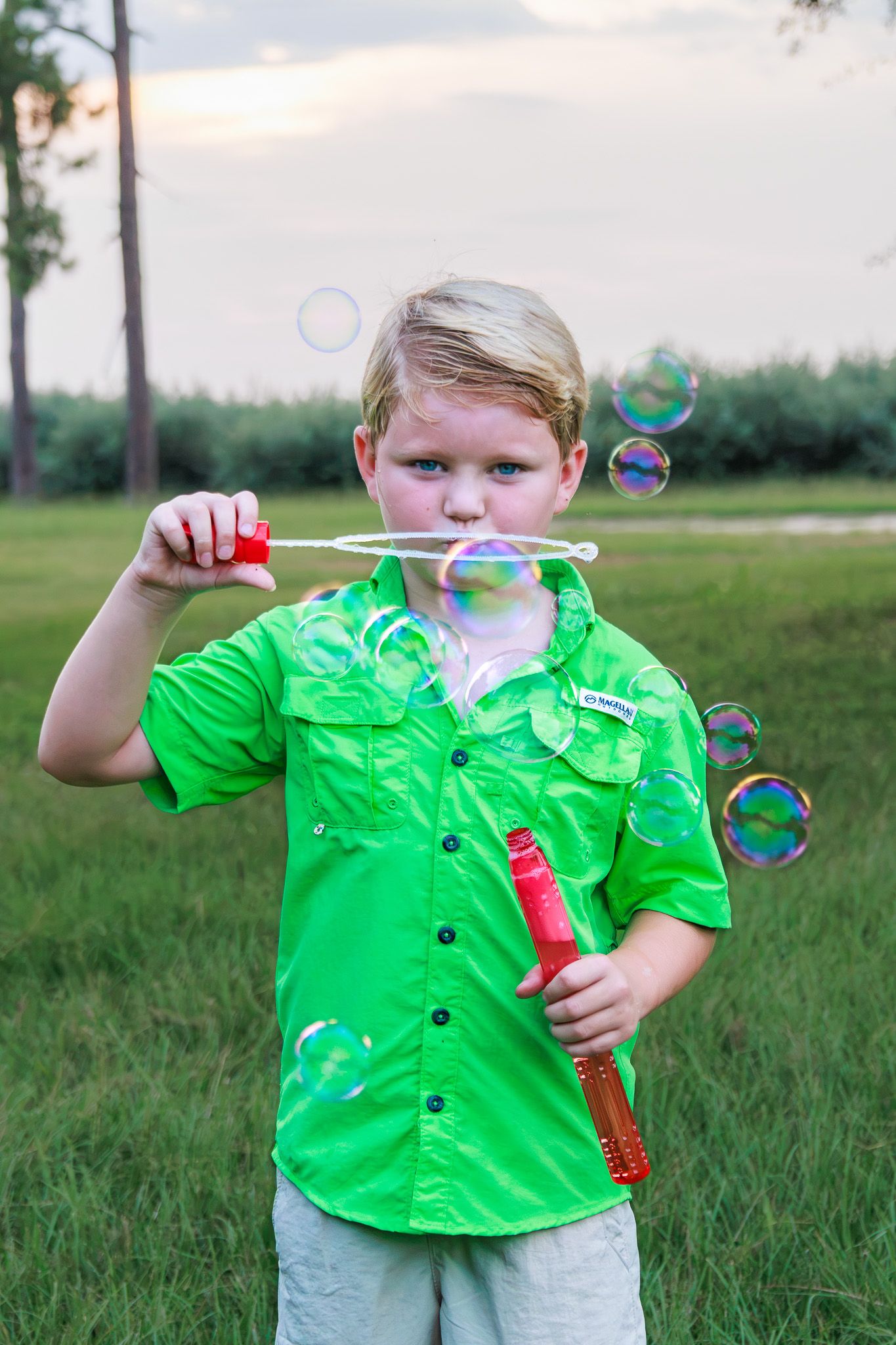 A young blonde boy in a green shirt blows colorful soap bubbles outdoors in a grassy field at dusk.