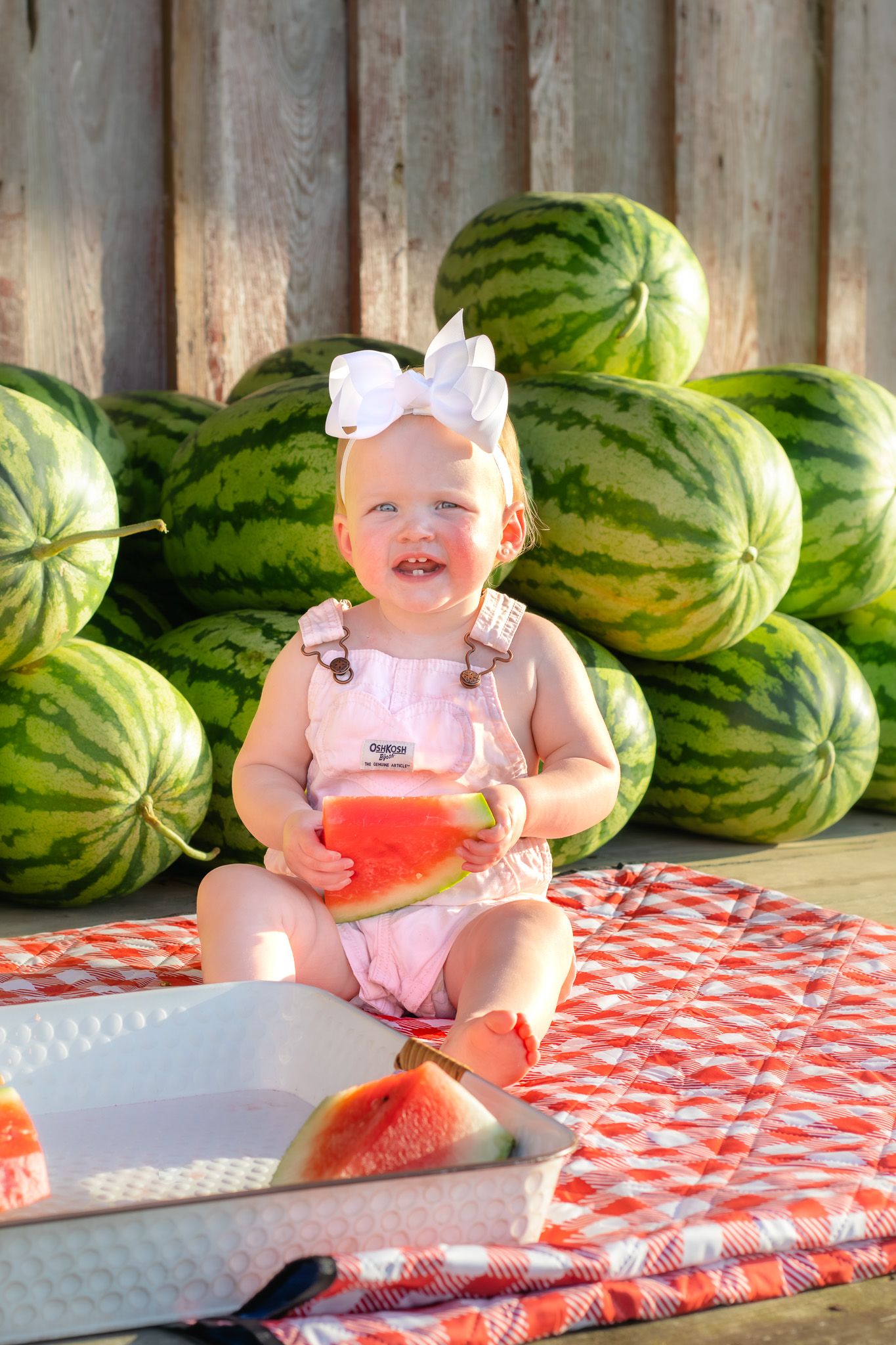 Smiling baby girl in pink overalls holding watermelon slice, sitting on checkered blanket surrounded by watermelons.