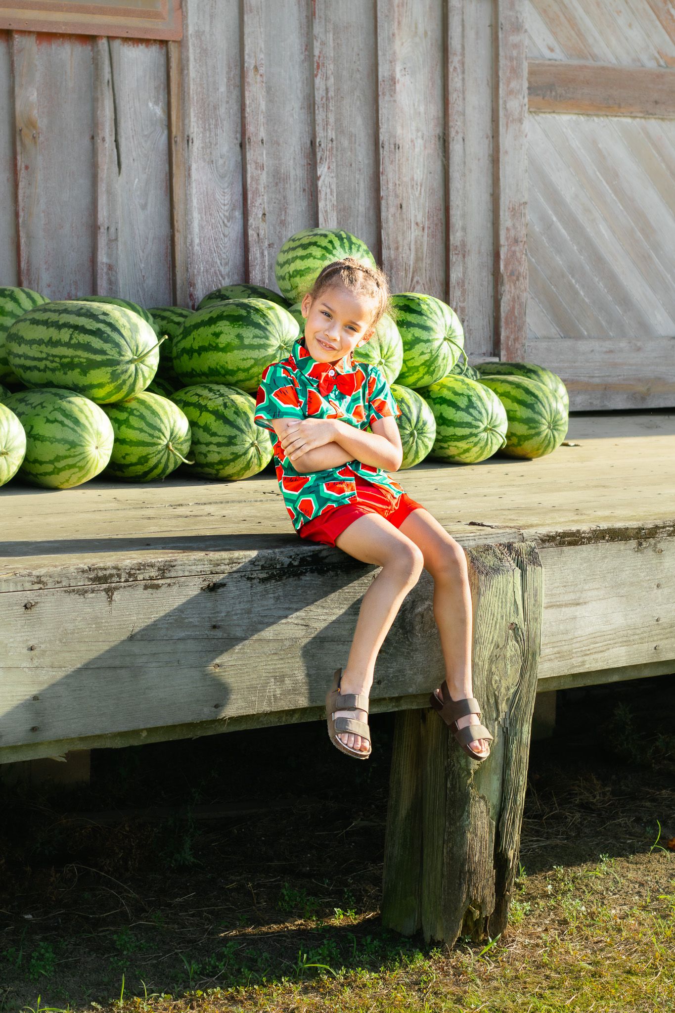 Young boy in watermelon shirt and red shorts sits on wooden platform with watermelons behind him.