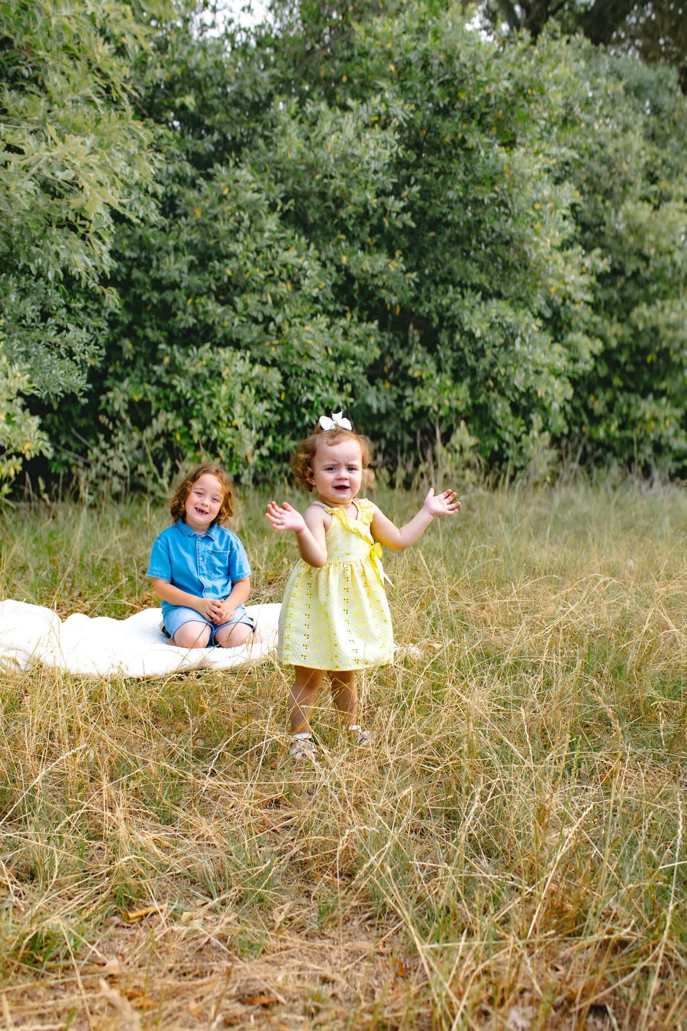 Two young children playing outdoors in a grassy field, girl in yellow dress with arms outstretched smiling joyfully.