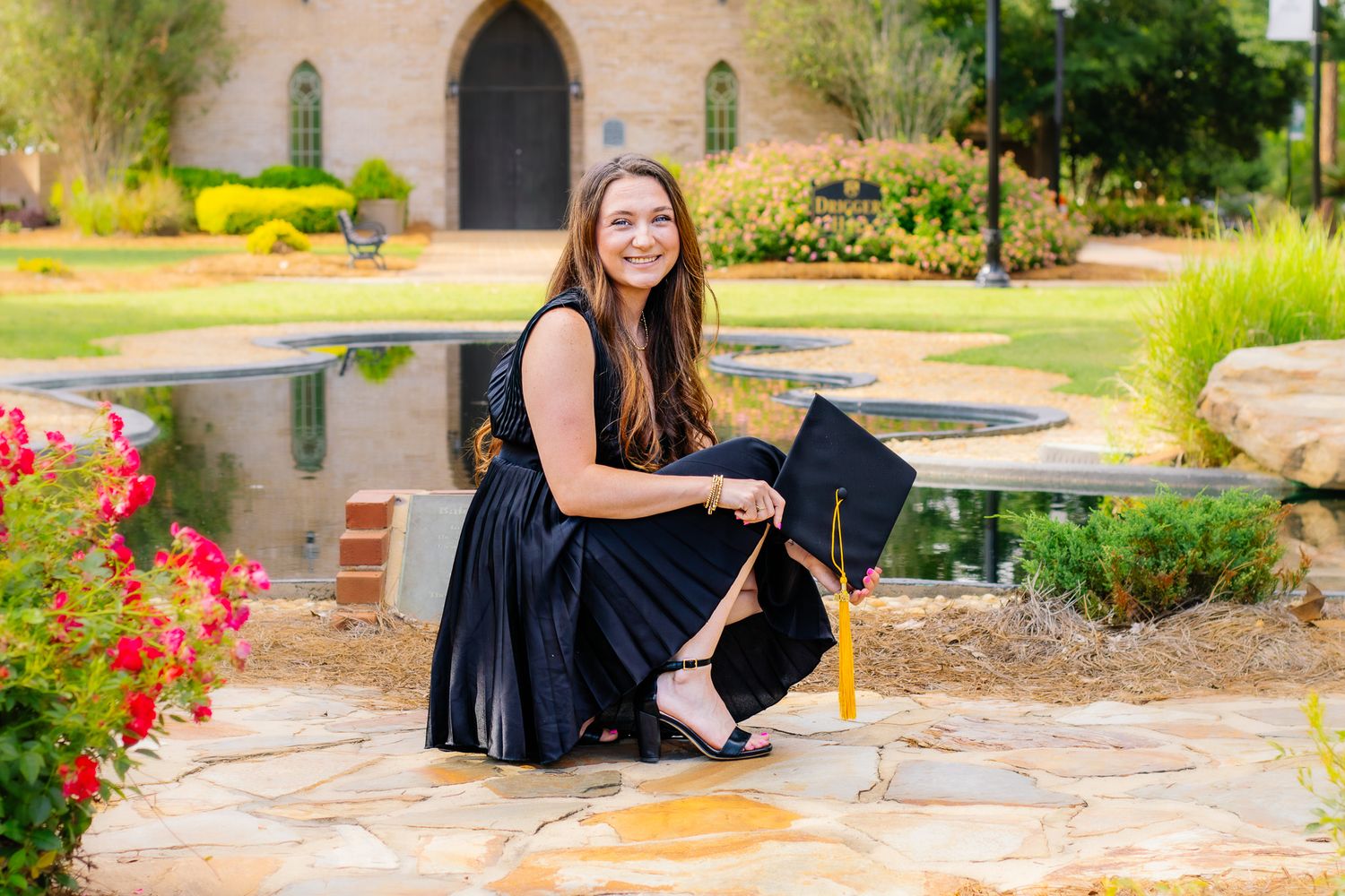 Smiling female graduate crouches on stone path holding cap near reflective pond and colorful flowers.