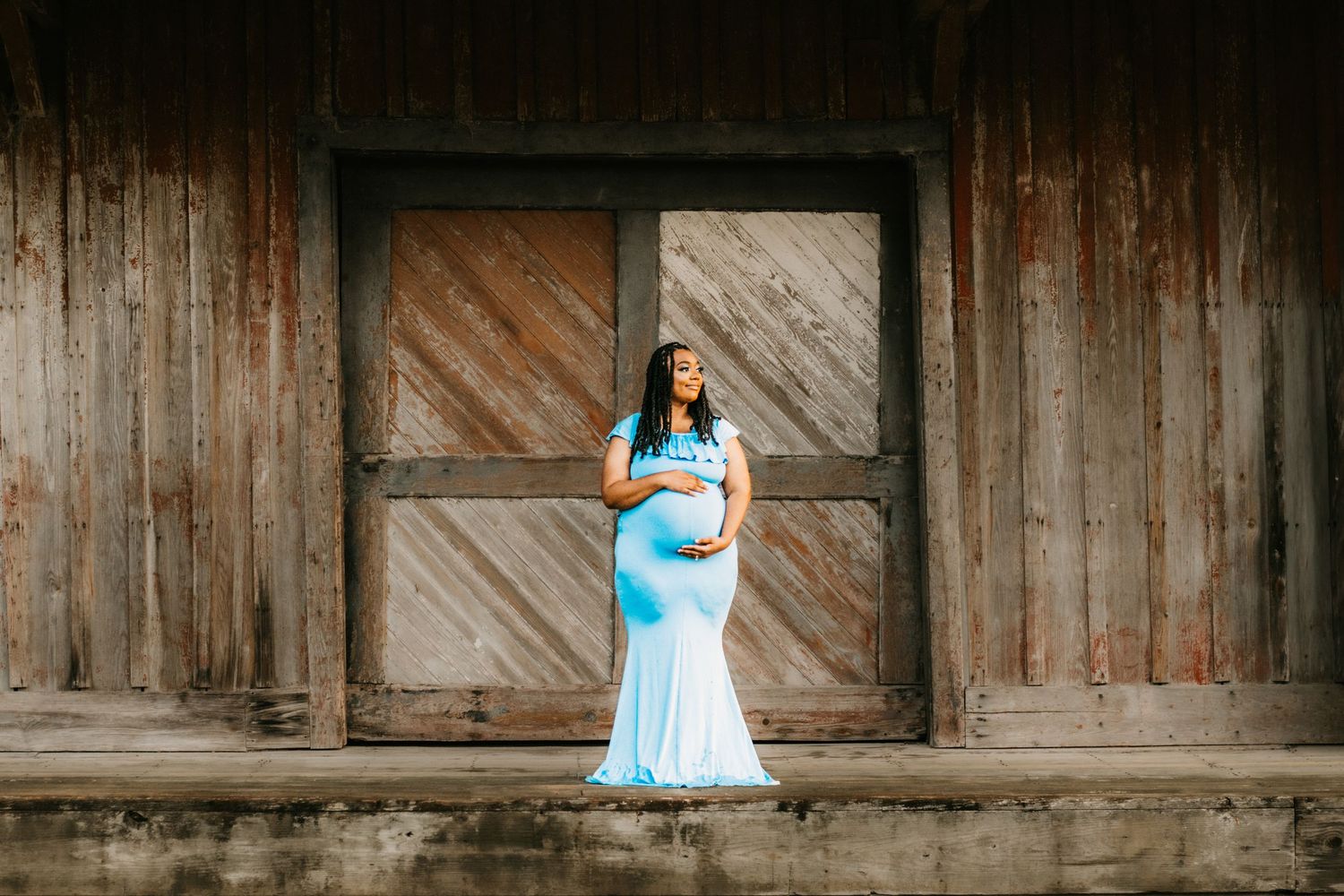 Pregnant woman in light blue mermaid gown poses gracefully in front of rustic wooden barn doors.