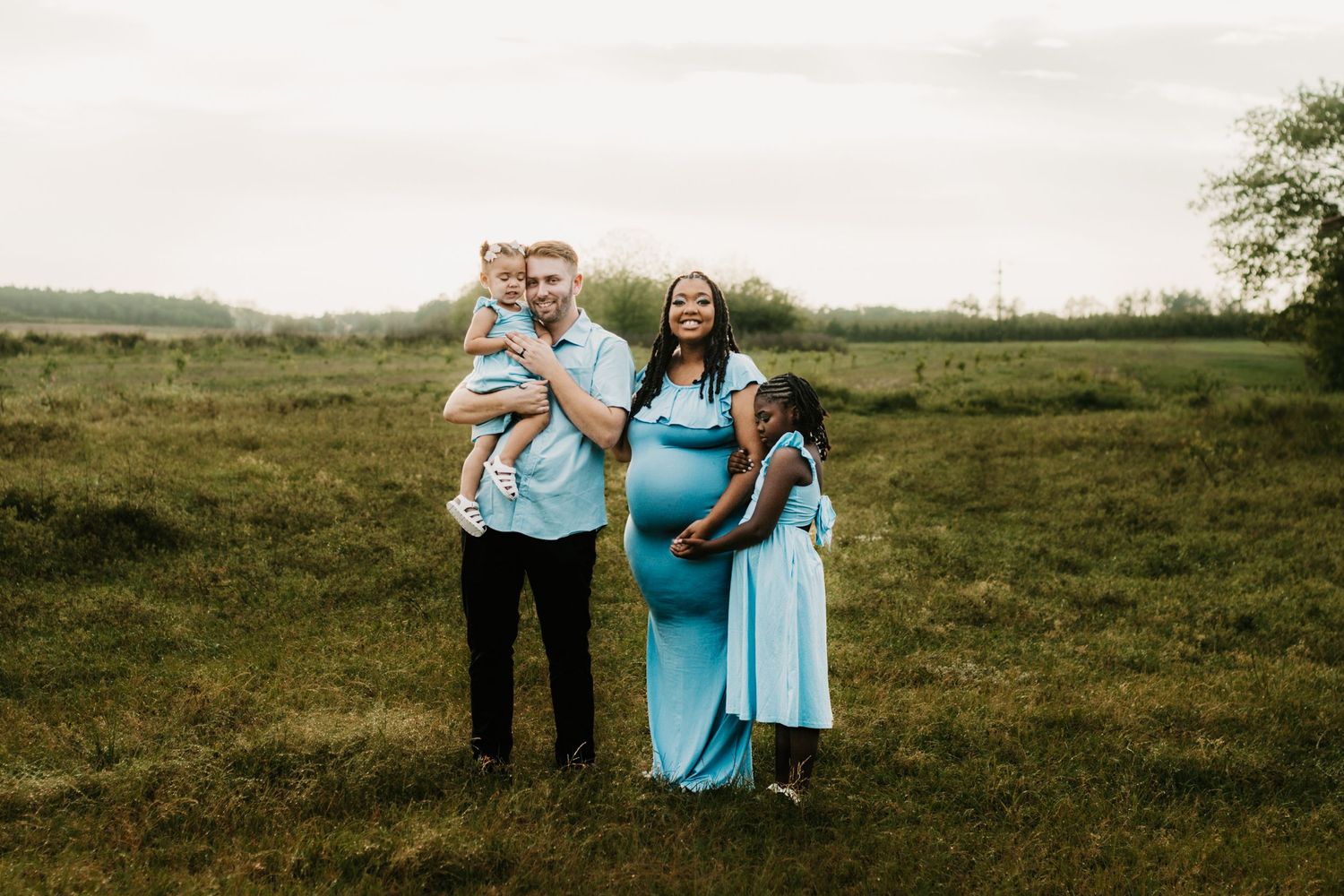 Smiling interracial family with two young children poses together in lush green field at sunset.