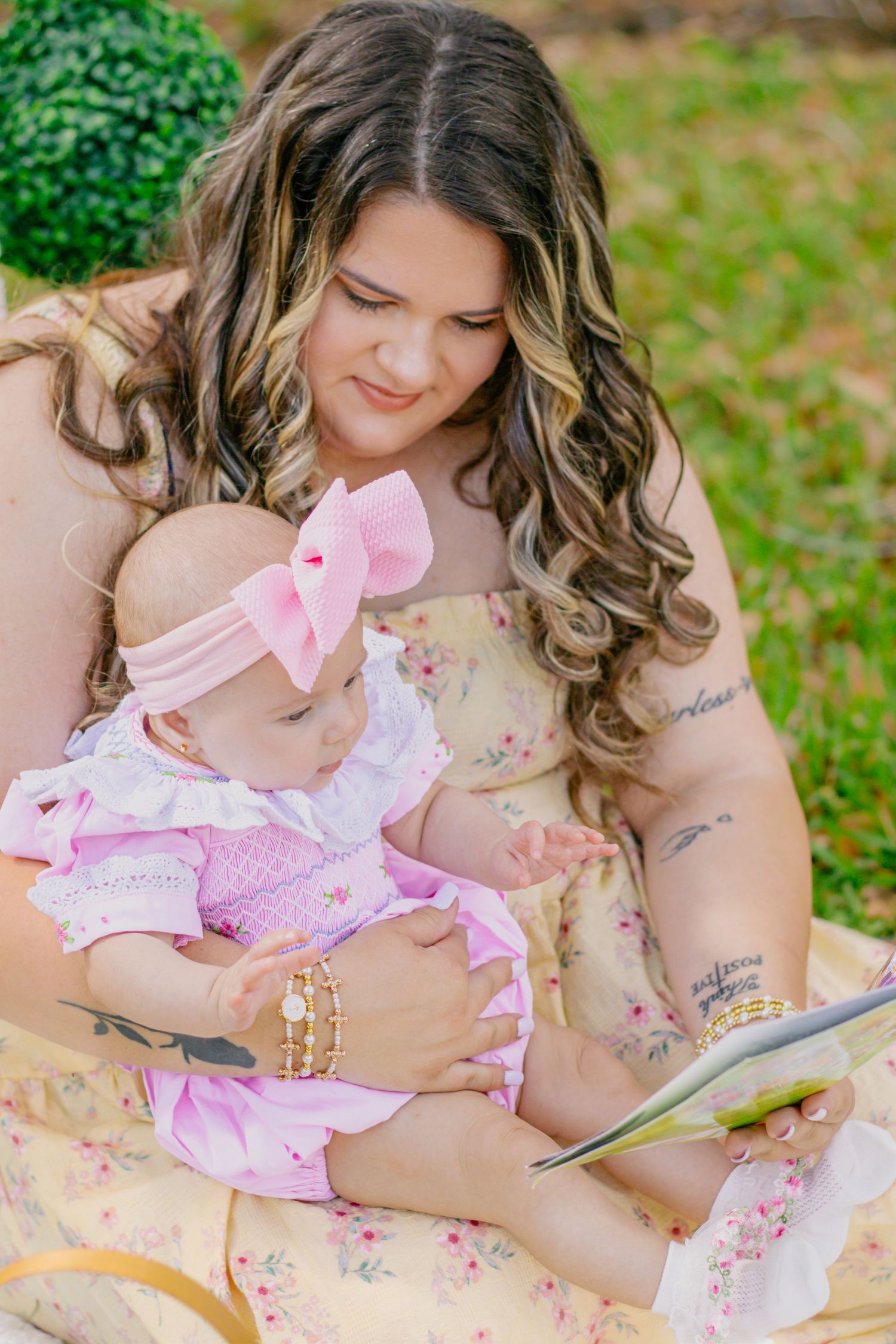 Mother in floral dress reading a book to her baby girl in pink outfit on outdoor blanket.