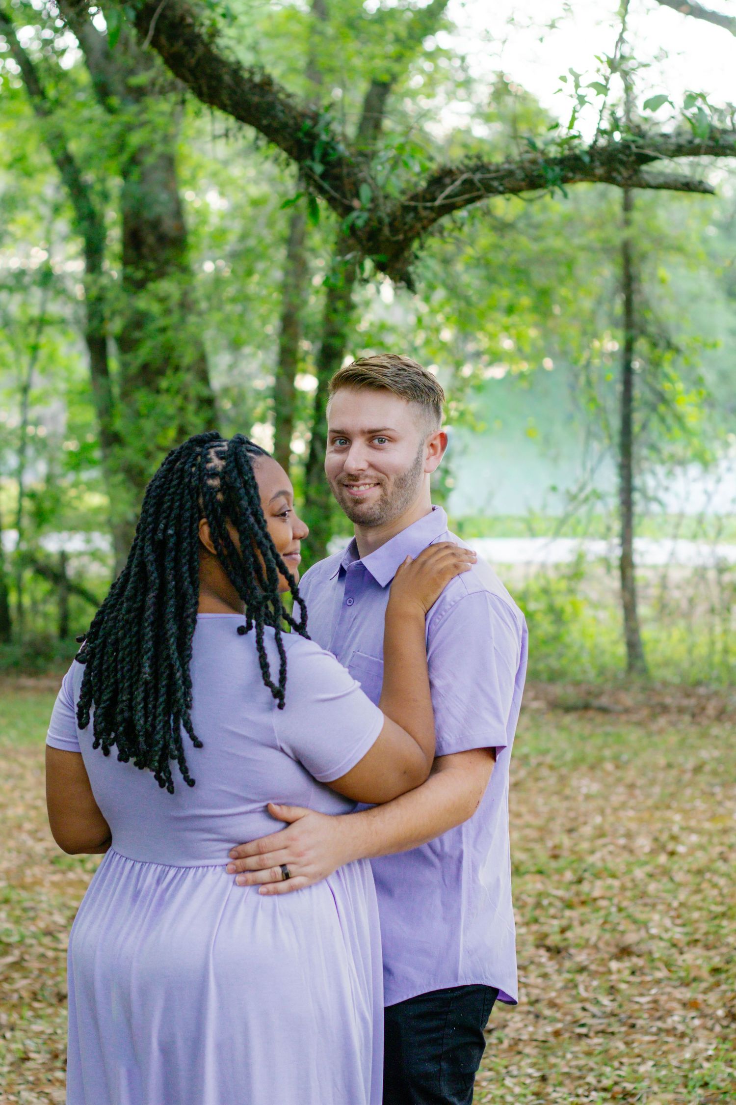 Interracial couple in coordinating lavender outfits share a tender embrace near a serene lake surrounded by trees.