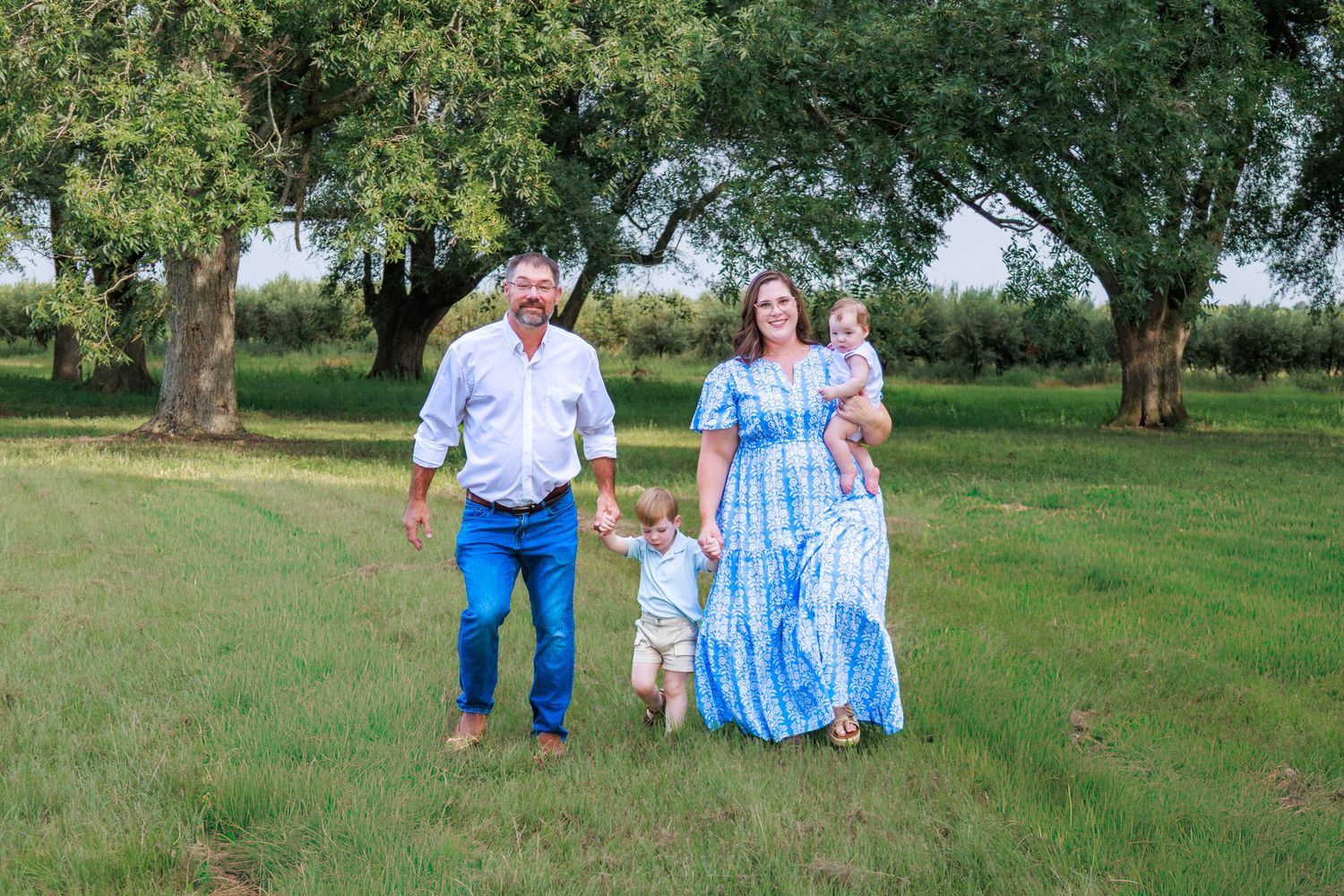 Family of four walking in green field with trees, parents holding children in blue outfits on sunny day.