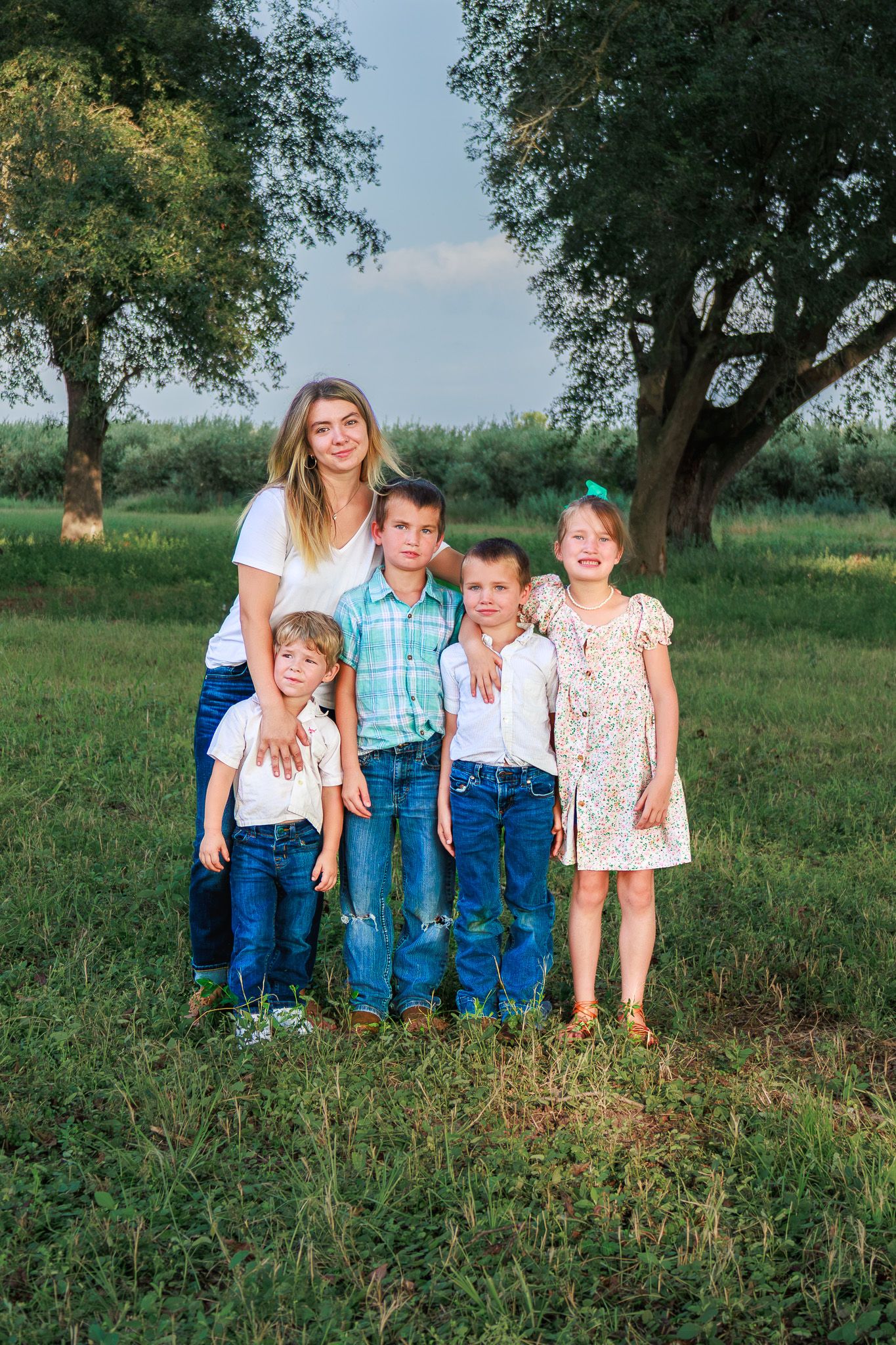 A woman poses outdoors with four children on green grass between large trees on a sunny day.