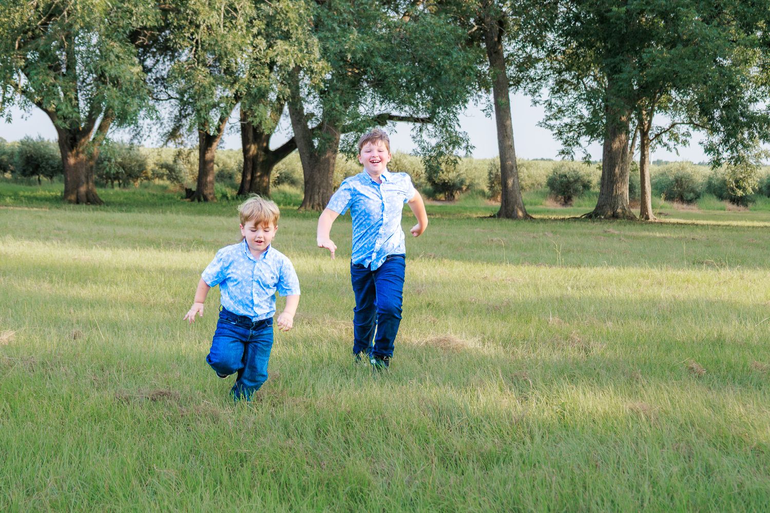 Two young boys in blue shirts and navy pants running joyfully through a lush green field with trees.