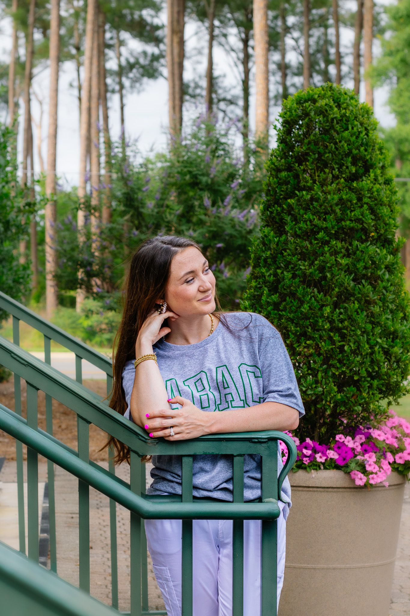 Woman in ABAC gray shirt leans on green railing surrounded by lush greenery and pink flowers.