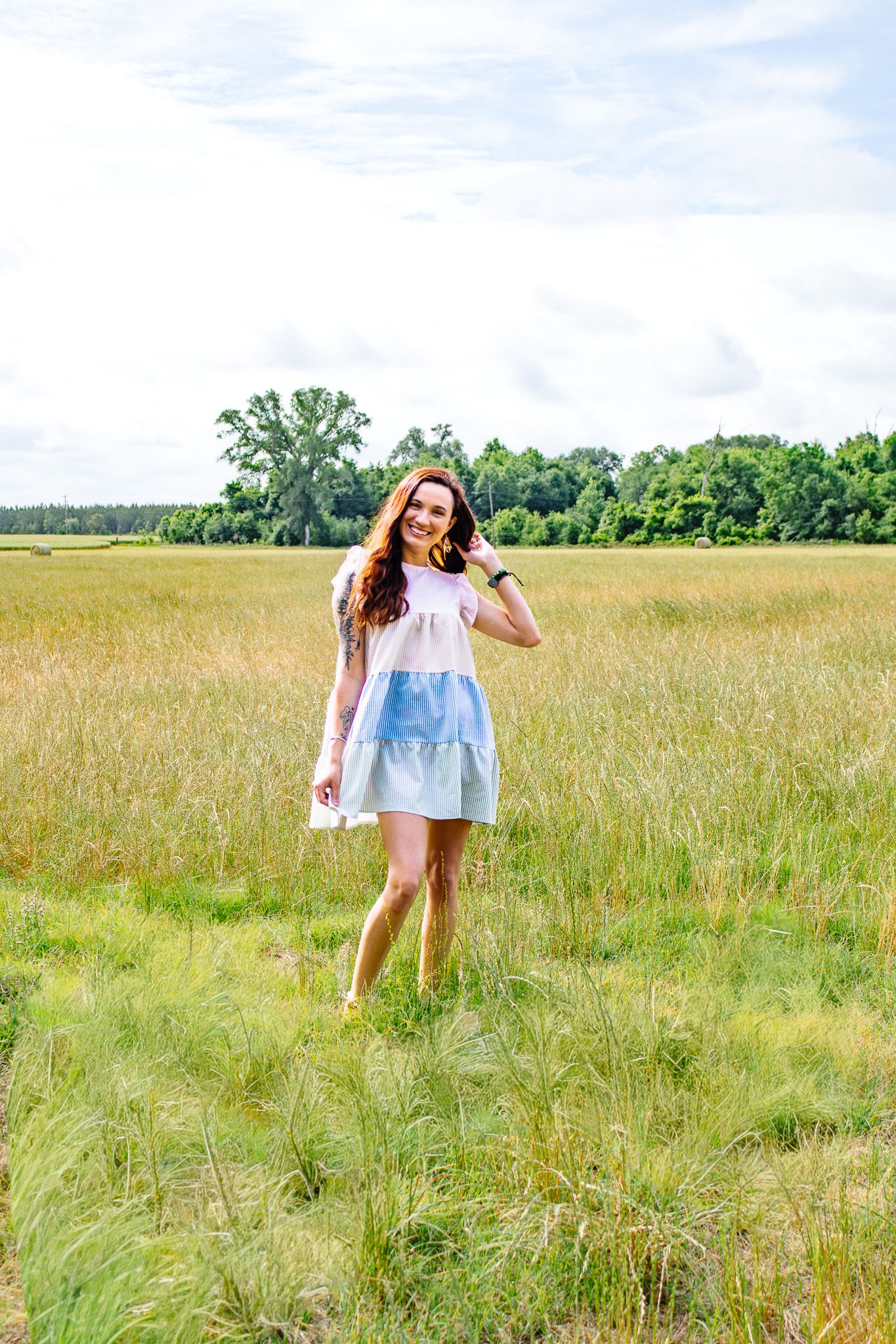 Young woman in white and blue sundress smiling while standing in a golden grassy field on a cloudy day.