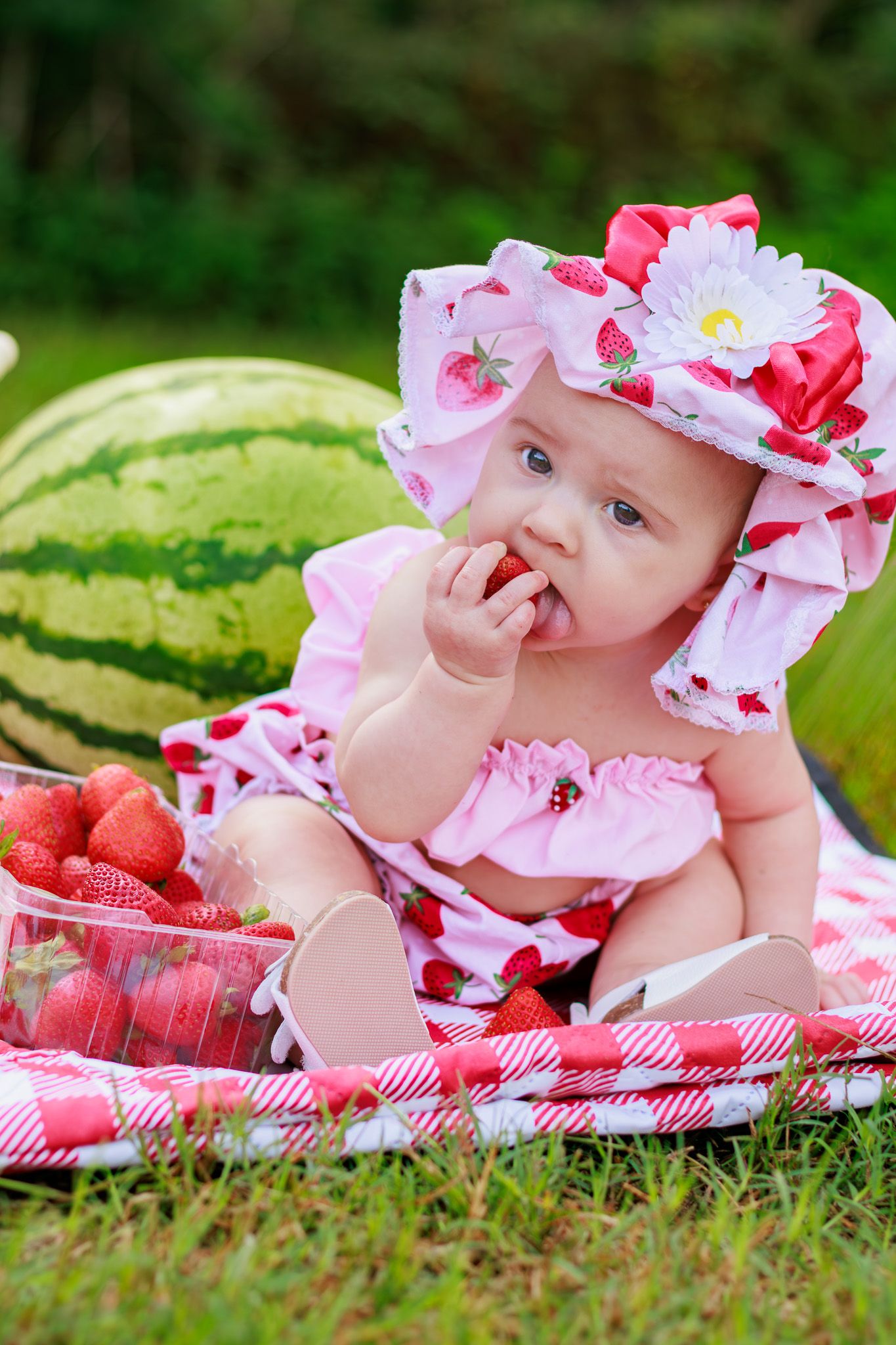 Adorable baby in strawberry print outfit eats fresh strawberry on picnic blanket beside large watermelon outdoors.