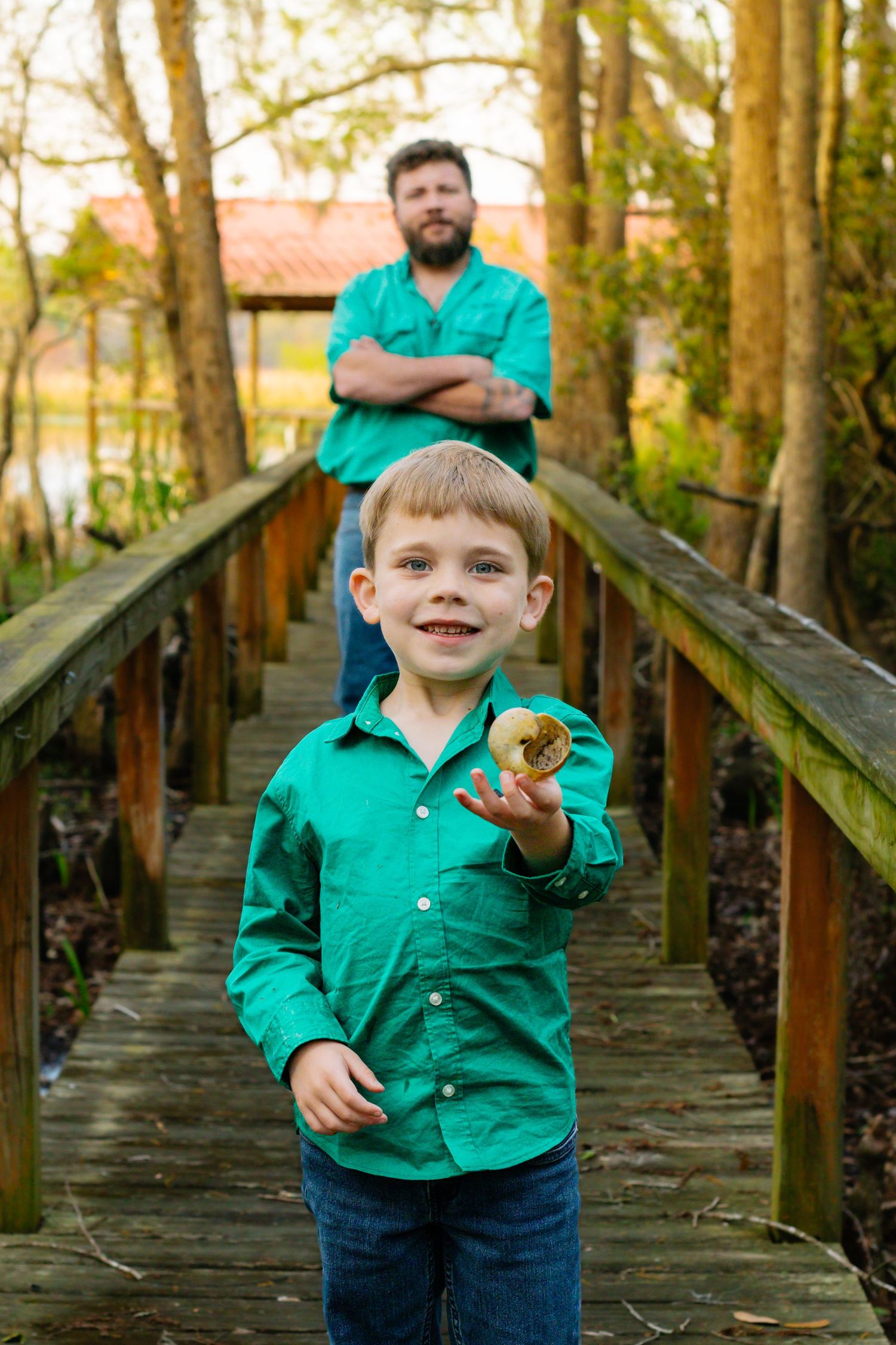 Young boy in green shirt holds object on wooden bridge with bearded father standing behind him.