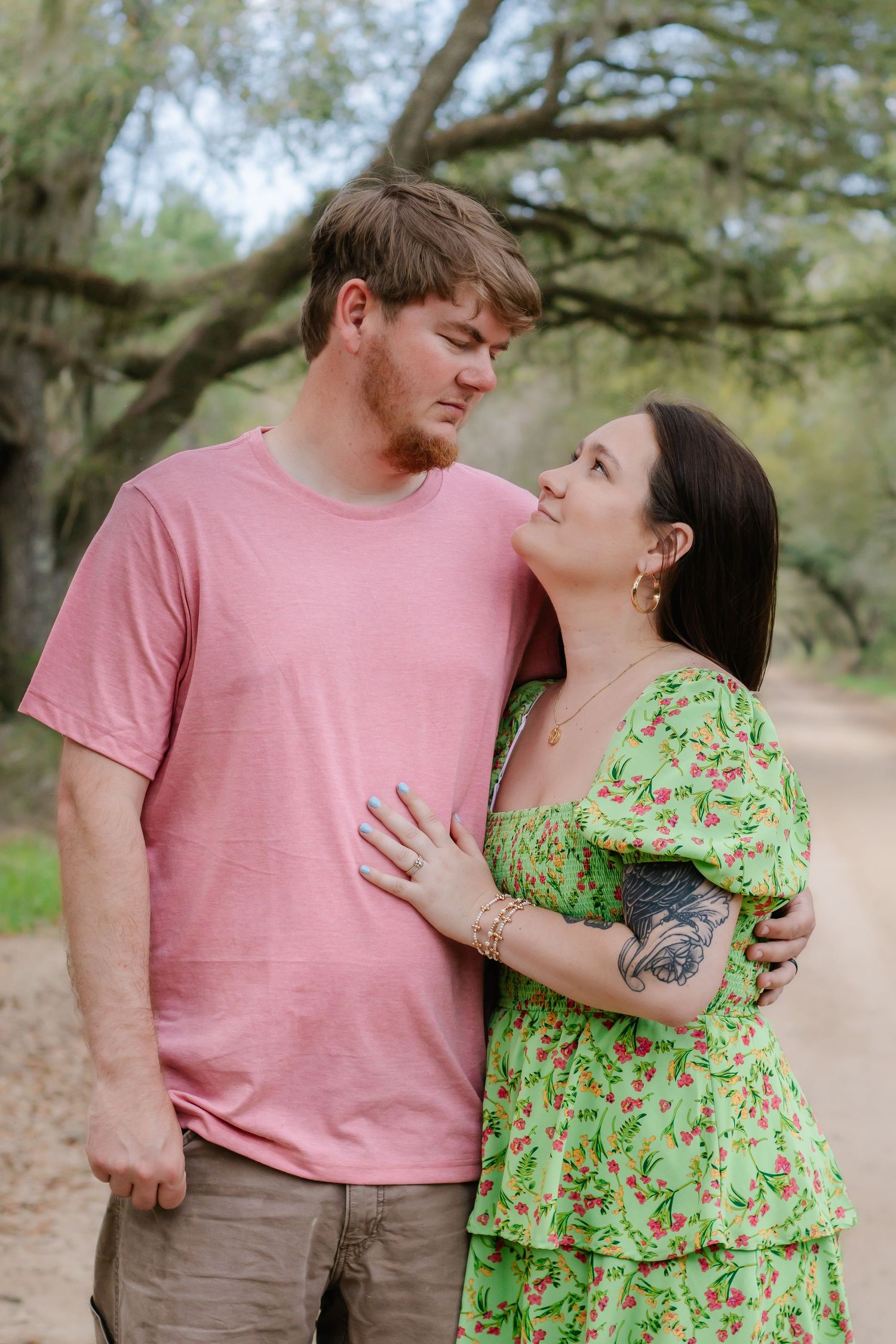 Romantic couple gazing at each other on a nature trail, woman in green floral dress embracing man in pink shirt.