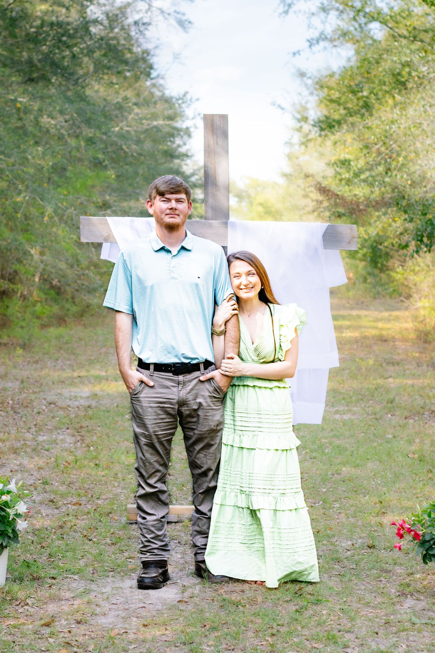 Young couple posing in front of rustic wooden cross outdoors, woman in green dress, man in blue polo.