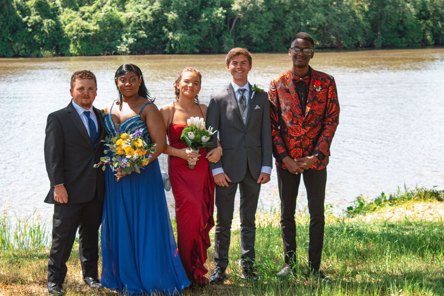 Five prom-dressed teens pose by a river, wearing blue, red, gray, and floral formal attire.