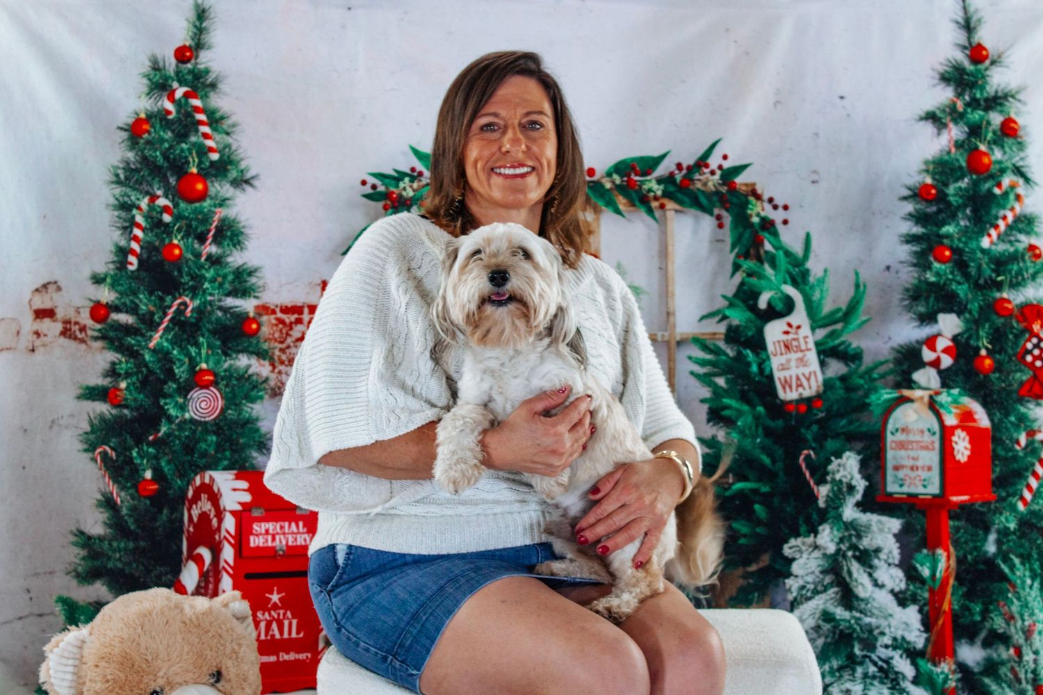 Smiling woman holds white fluffy dog in festive Christmas photo session with decorated trees.