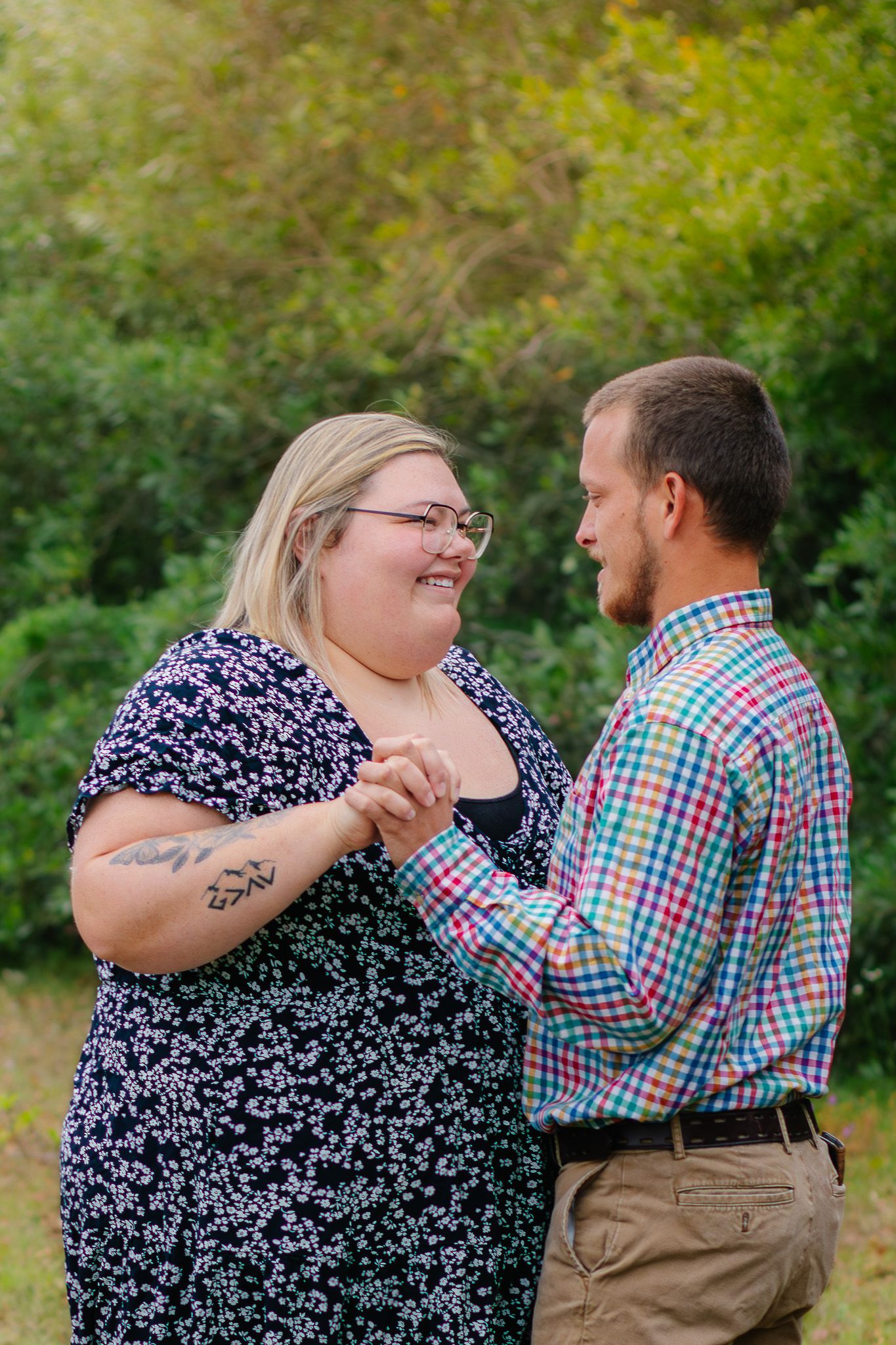 A couple holds hands and smiles at each other outdoors, woman in floral dress with tattoos, man in plaid shirt.