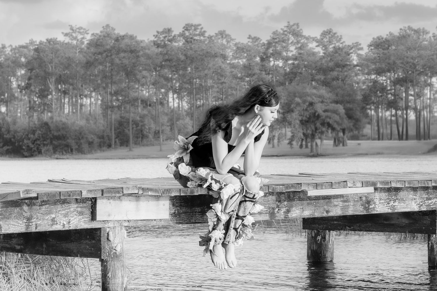 Black and white photo of a woman in a floral dress sitting pensively on a wooden dock by a lake.
